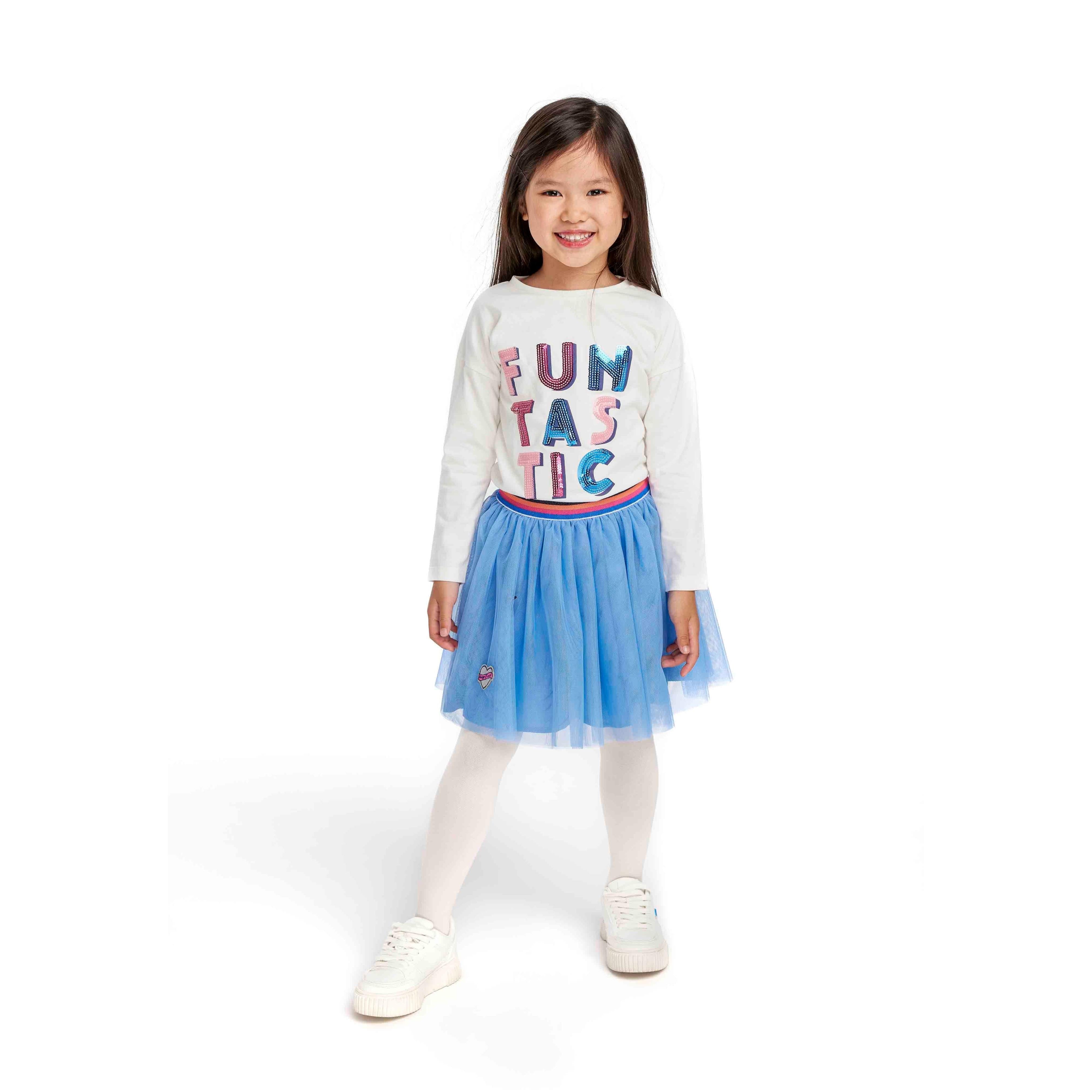 Young girl wearing cream long sleeve t-shirt with sequin FUNTASTIC lettering and blue tulle skirt, white tights and sneakers, smiling on white background