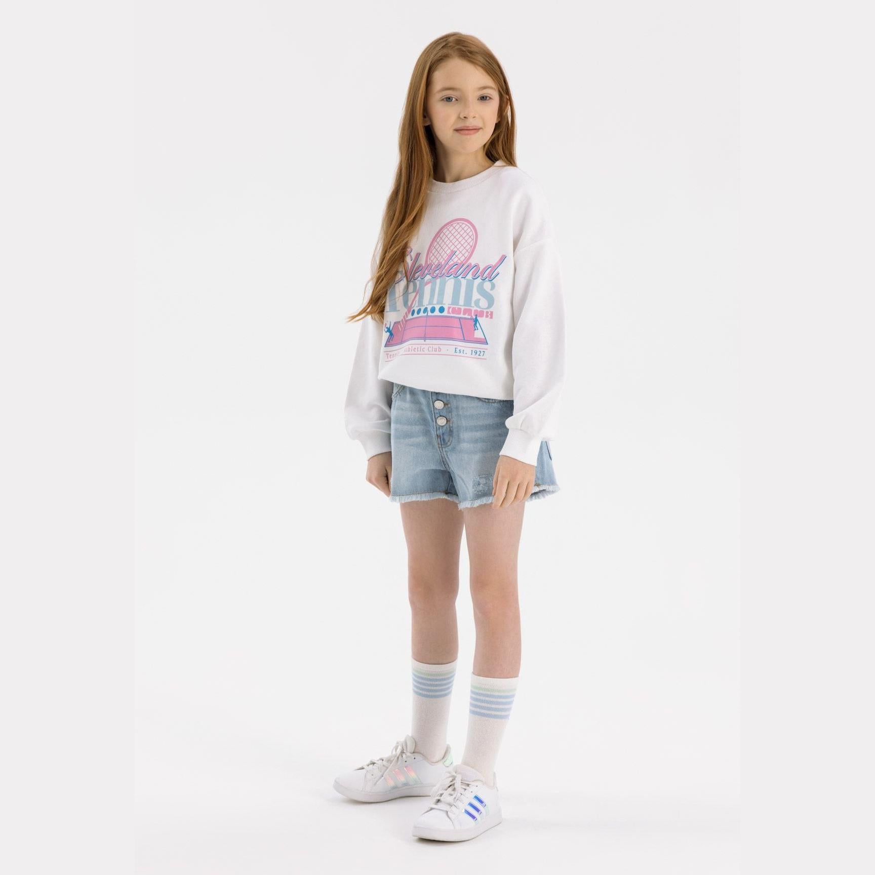 Young girl wearing light blue denim shorts with frayed hem and white sweatshirt with tennis racket graphic, standing against white background