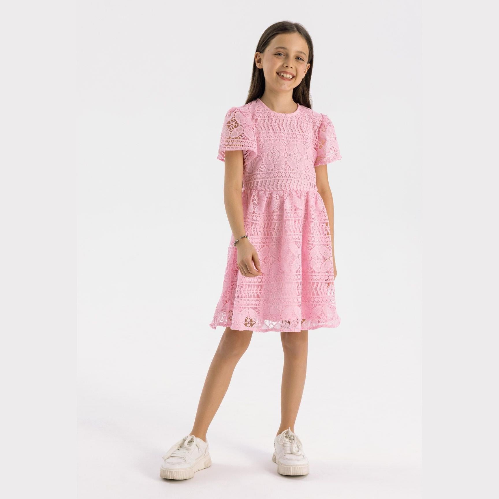 Young girl wearing pink lace dress with short sleeves, standing against white background, smiling and wearing white trainers