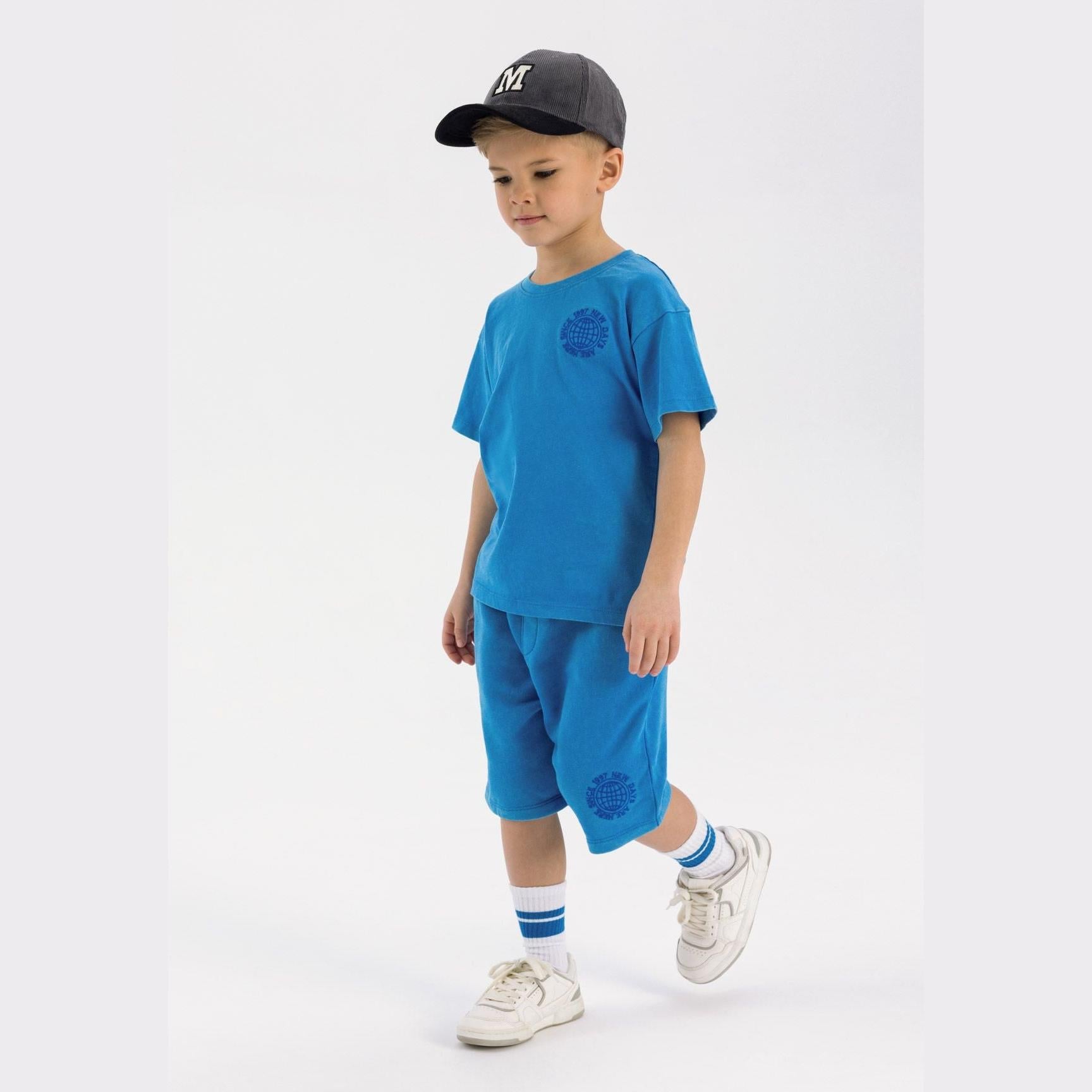 Young boy wearing blue t-shirt and shorts set with black NY cap, white sneakers and blue striped socks against white background