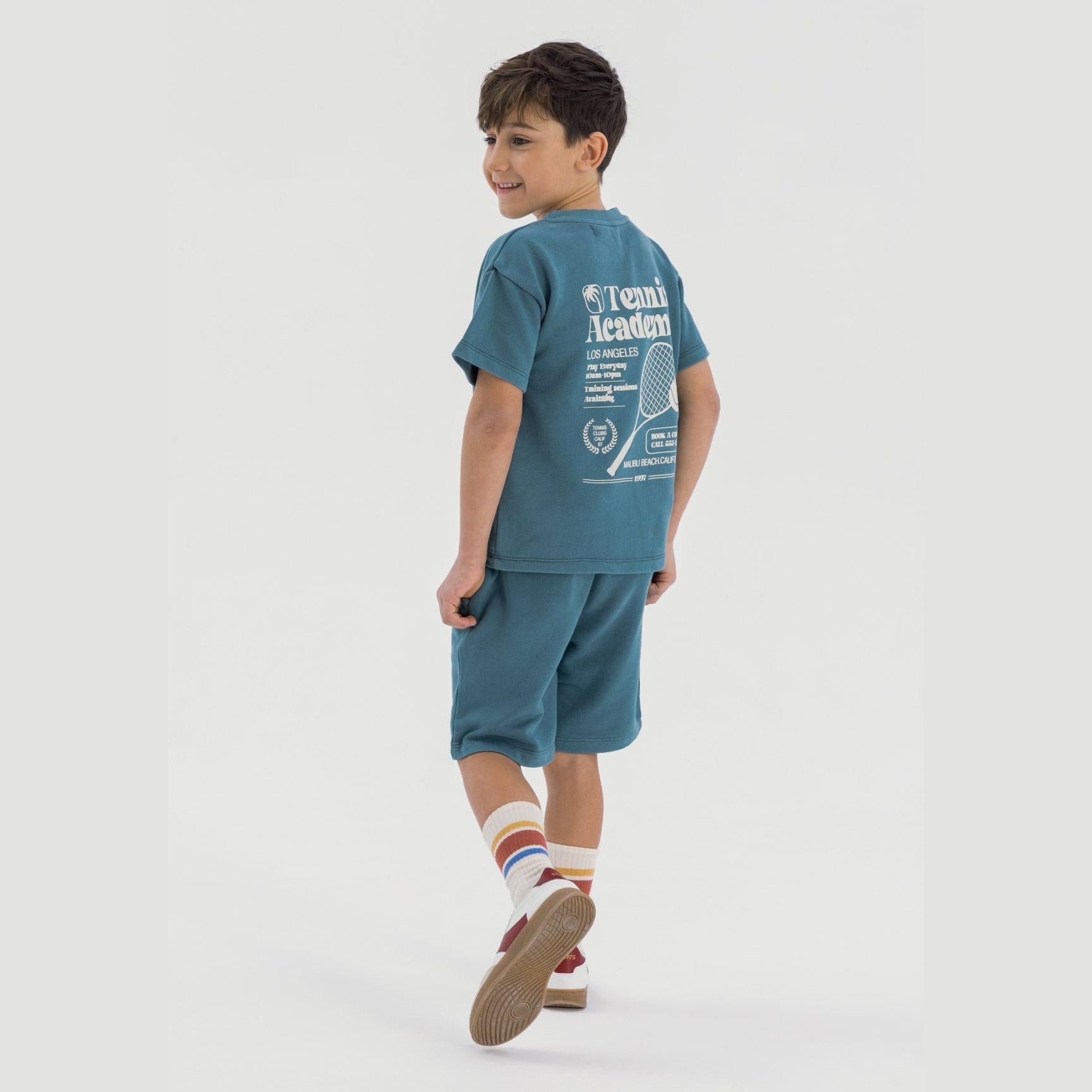 Young boy wearing teal tennis academy t-shirt and matching shorts, walking and smiling against white background, showing back view of tennis print