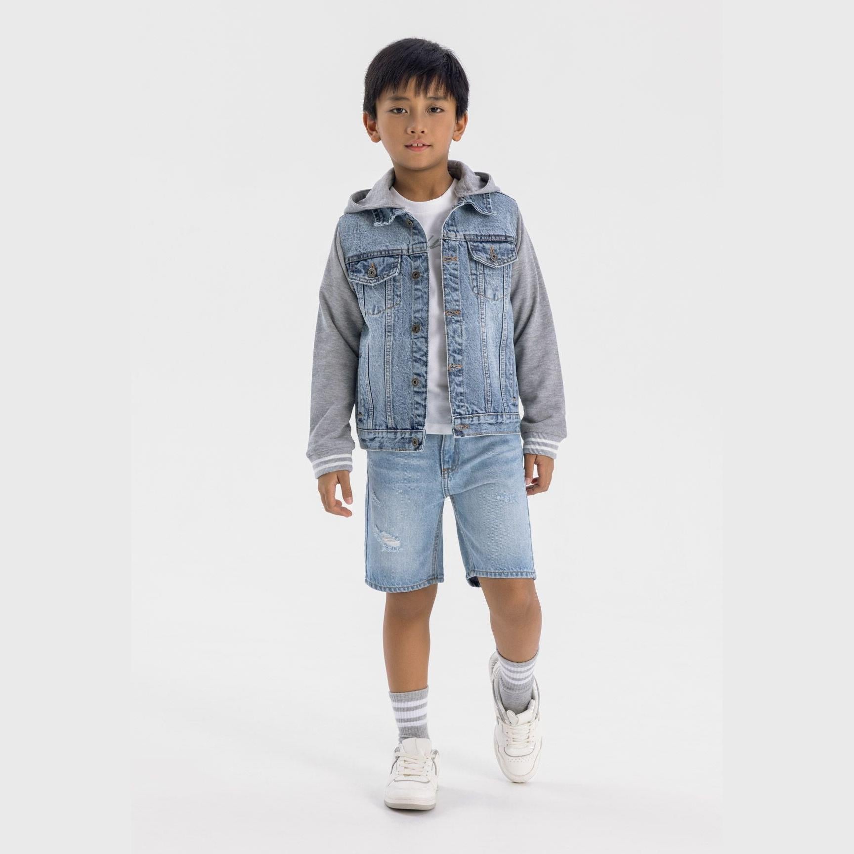 Young boy wearing light blue ripped denim shorts with denim jacket and grey hoodie, standing against white background