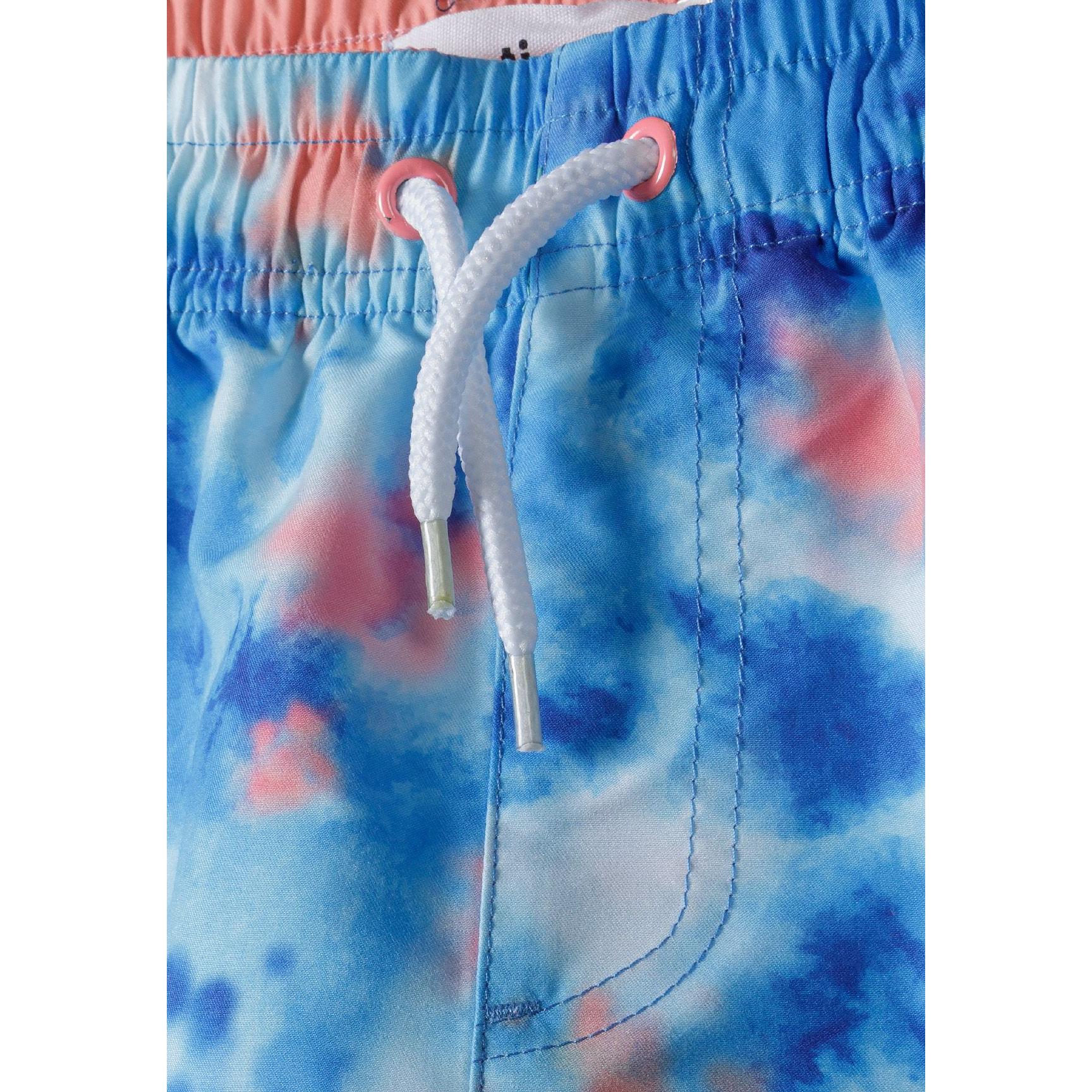 Close-up detail of blue tie-dye swim shorts showing white drawstring with coral eyelets and elastic waistband construction