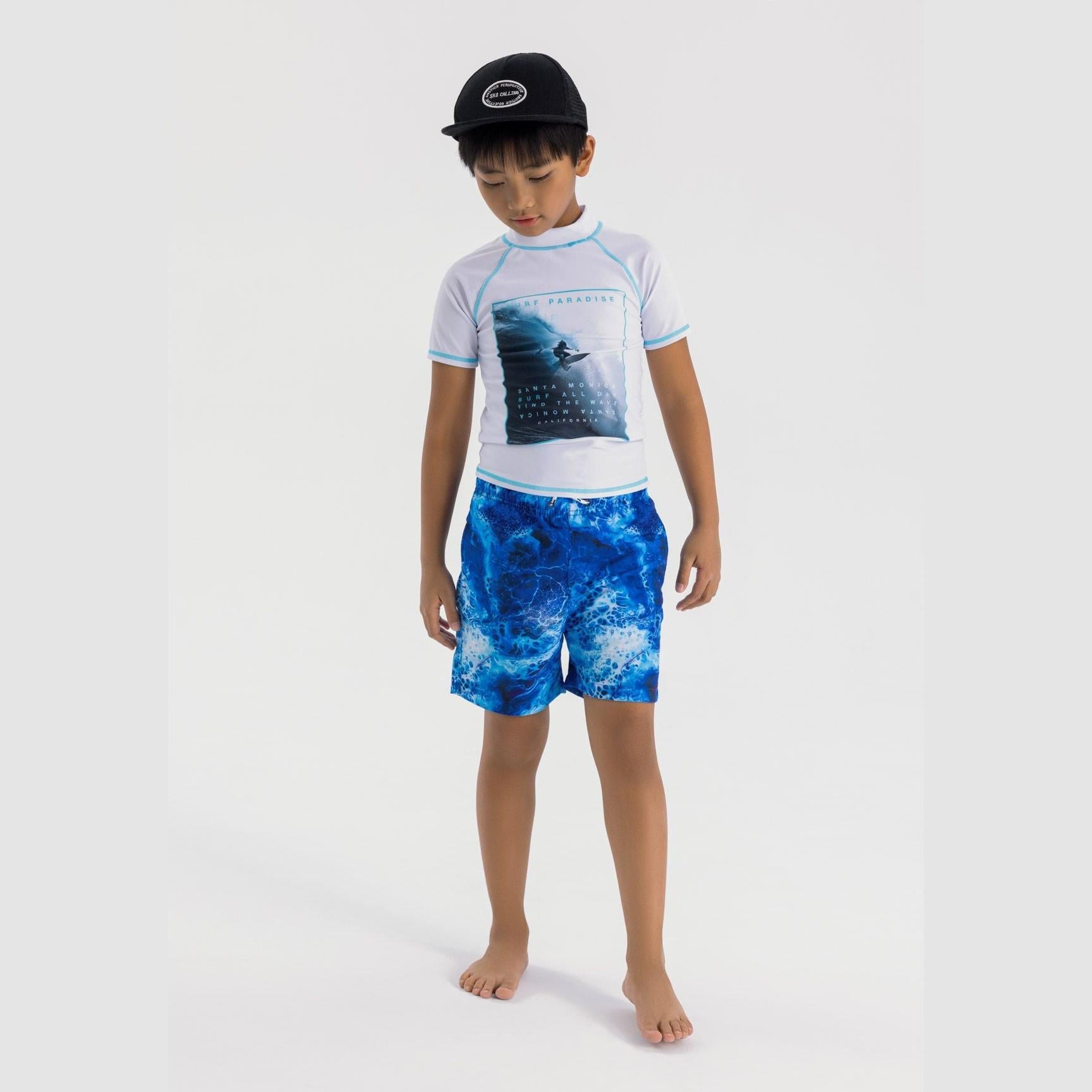 Young boy wearing white swim t-shirt with surf paradise print and blue patterned swim shorts, paired with black cap, standing barefoot against white background
