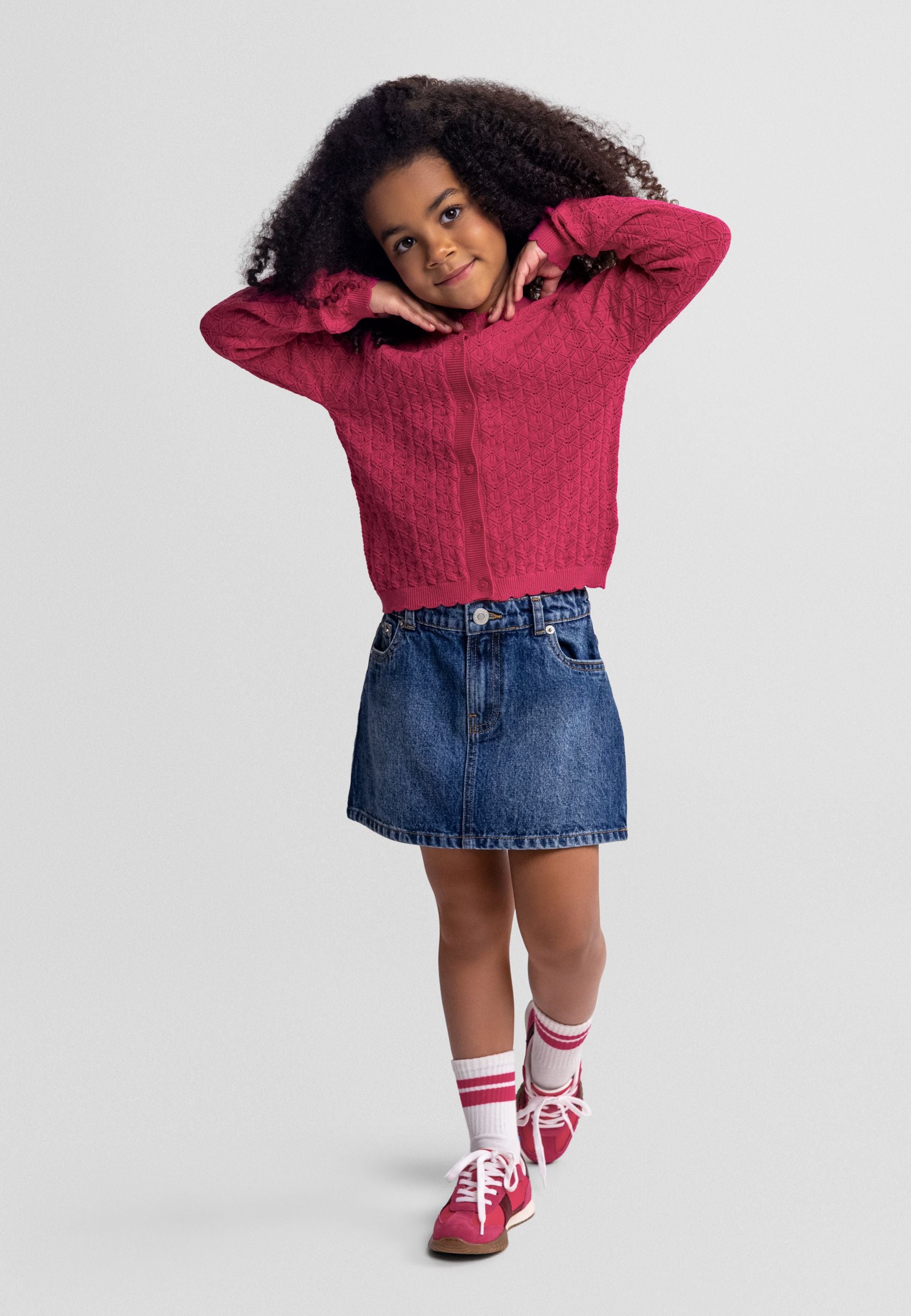 Young girl wearing dark blue denim A-line skirt with pink cable knit sweater, striped socks and red sneakers on white studio background