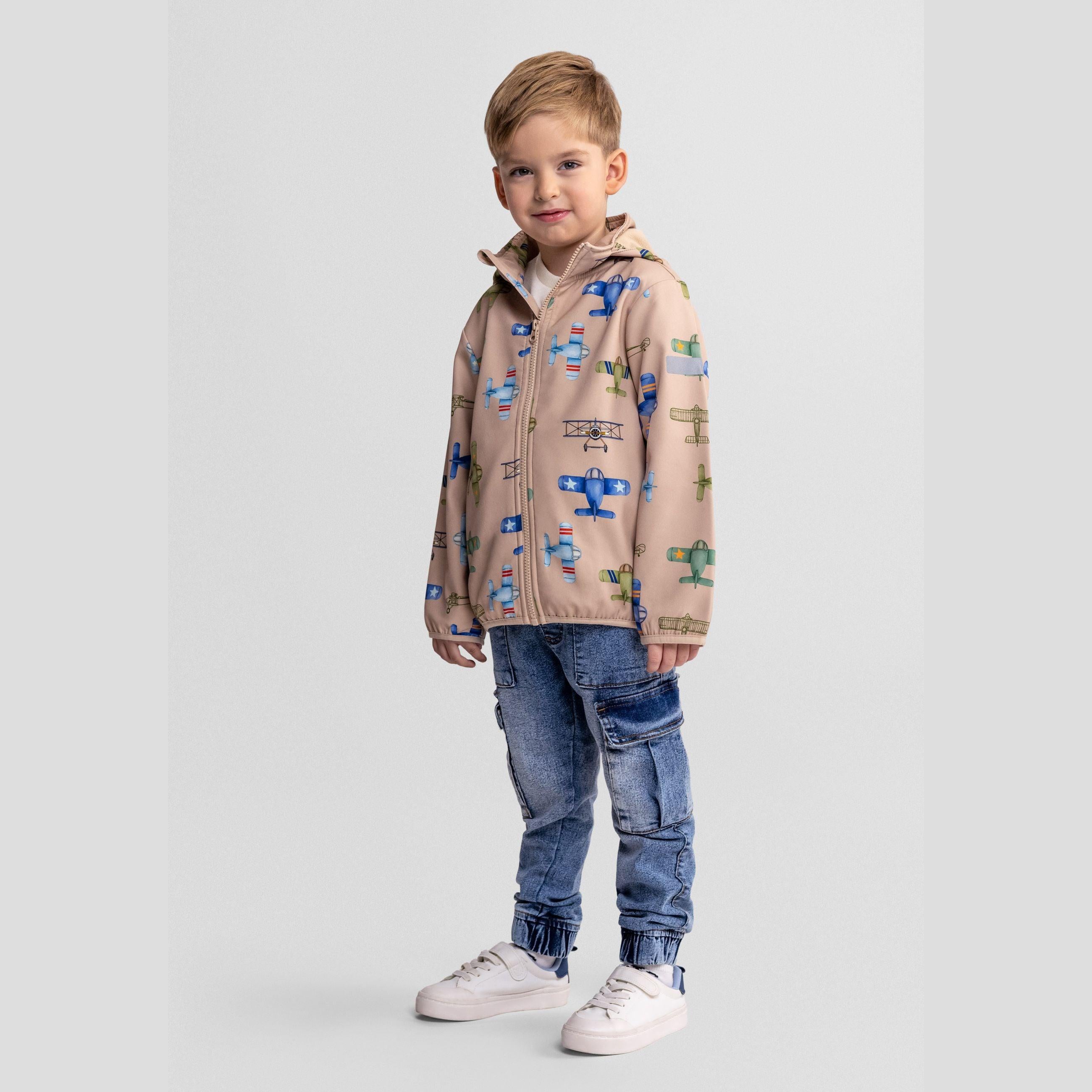 Young boy wearing sand-coloured softshell jacket with aeroplane print and hooded design, paired with distressed denim jeans and white trainers, standing against light grey background