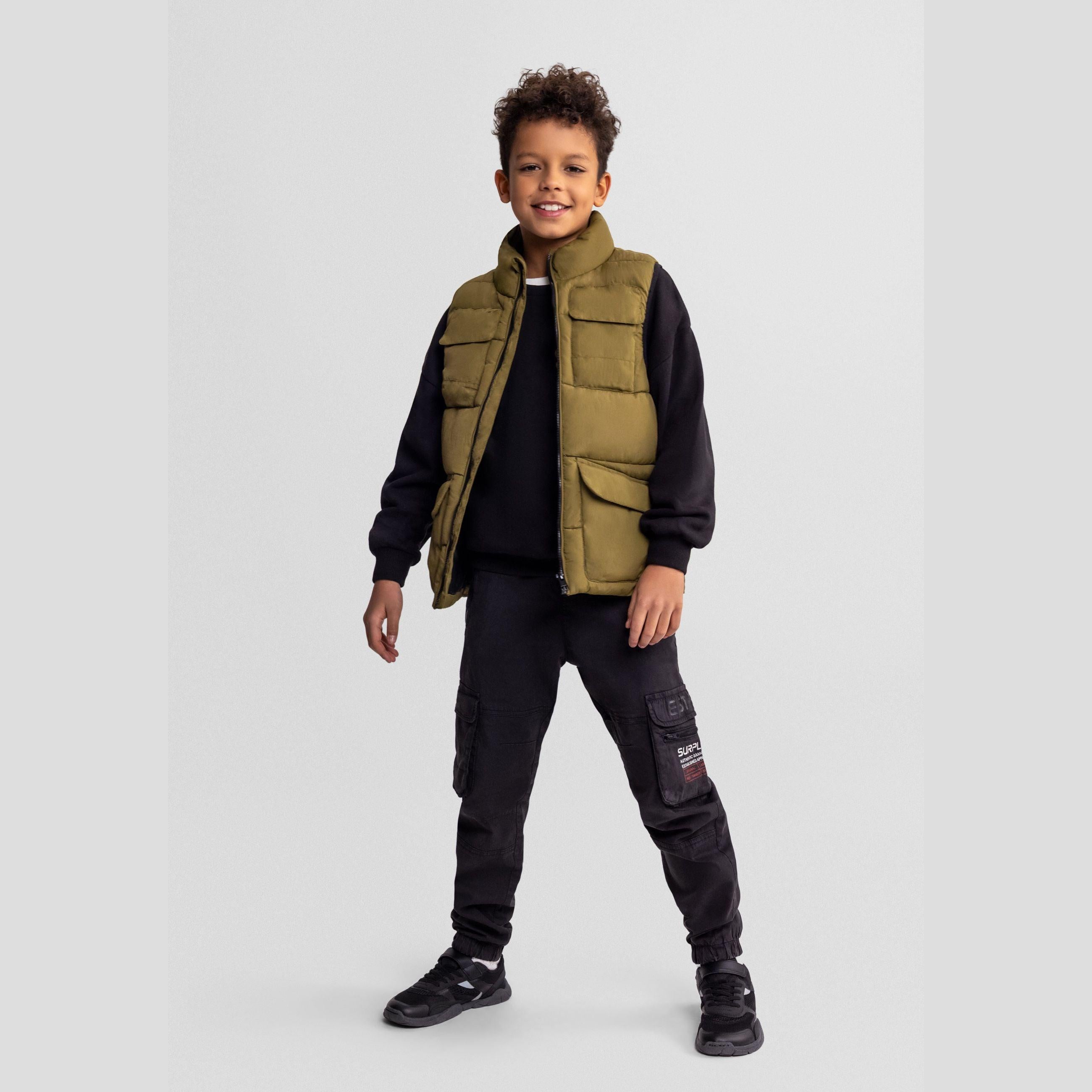 Young boy wearing olive green padded utility waistcoat with black sweatshirt and cargo pants, standing against grey background