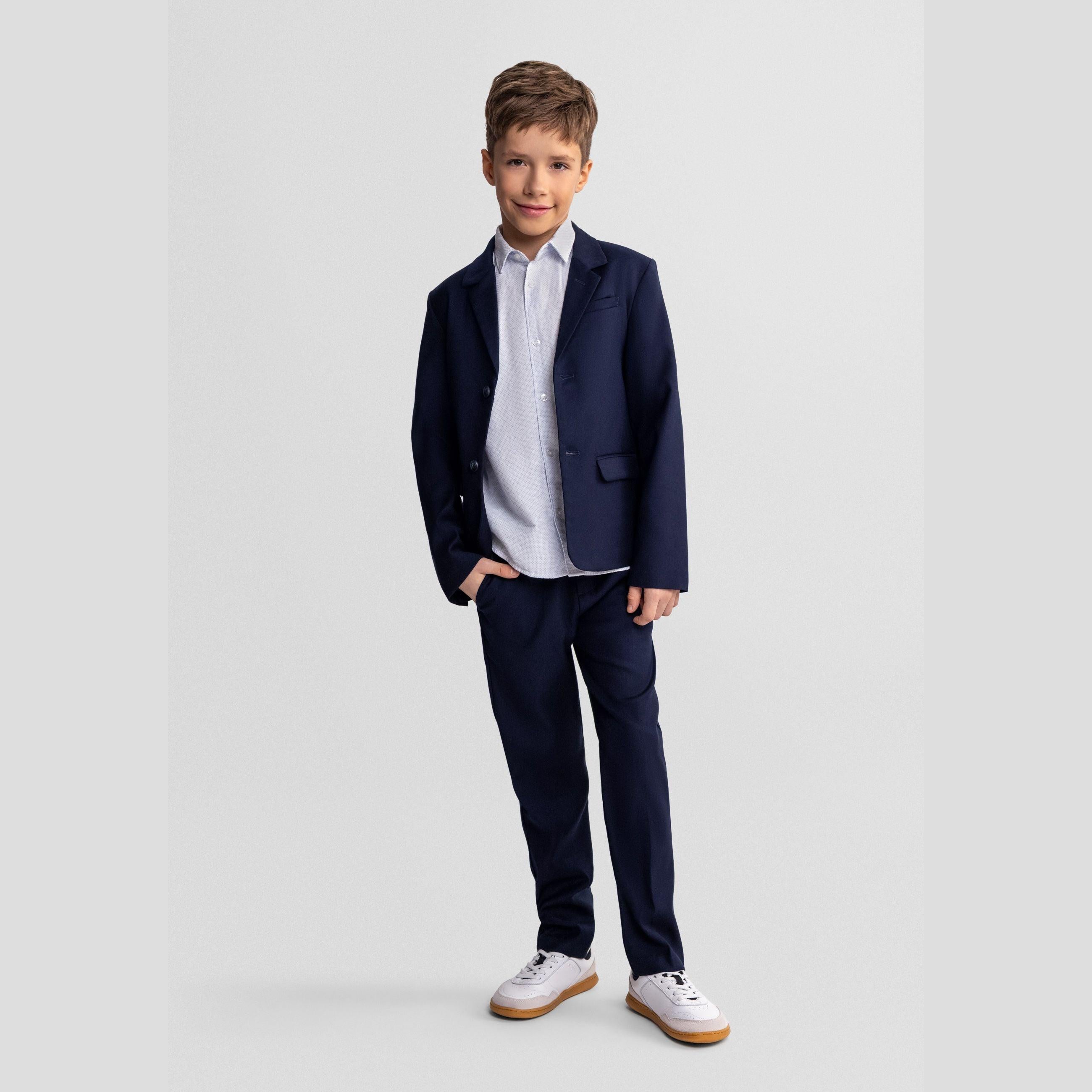 Boy wearing navy formal trousers with matching blazer and white shirt, standing against light grey studio background