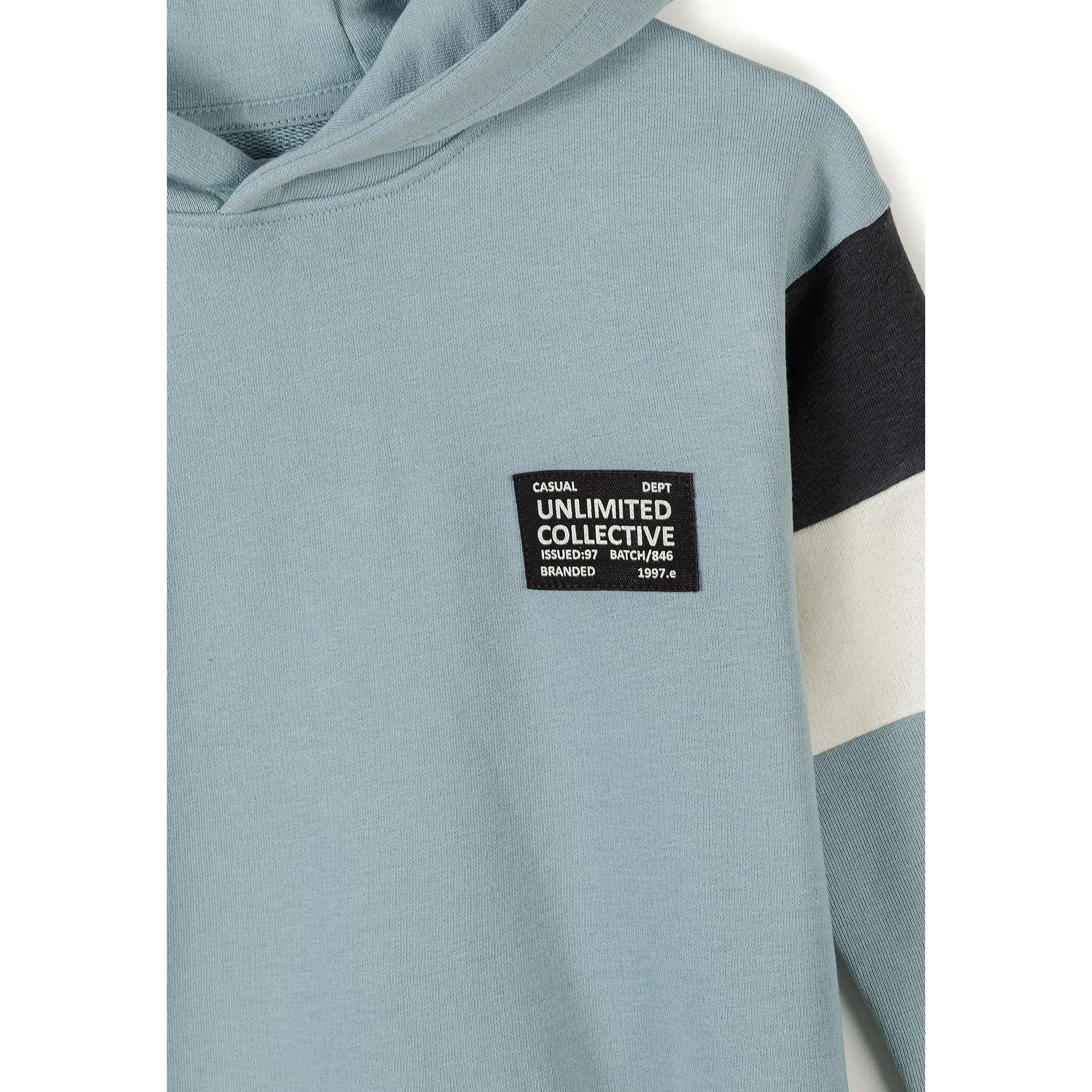 Close-up detail of sage blue jogger waistband and front pocket area showing black rectangular branded patch reading 'Unlimited Collective' with white text