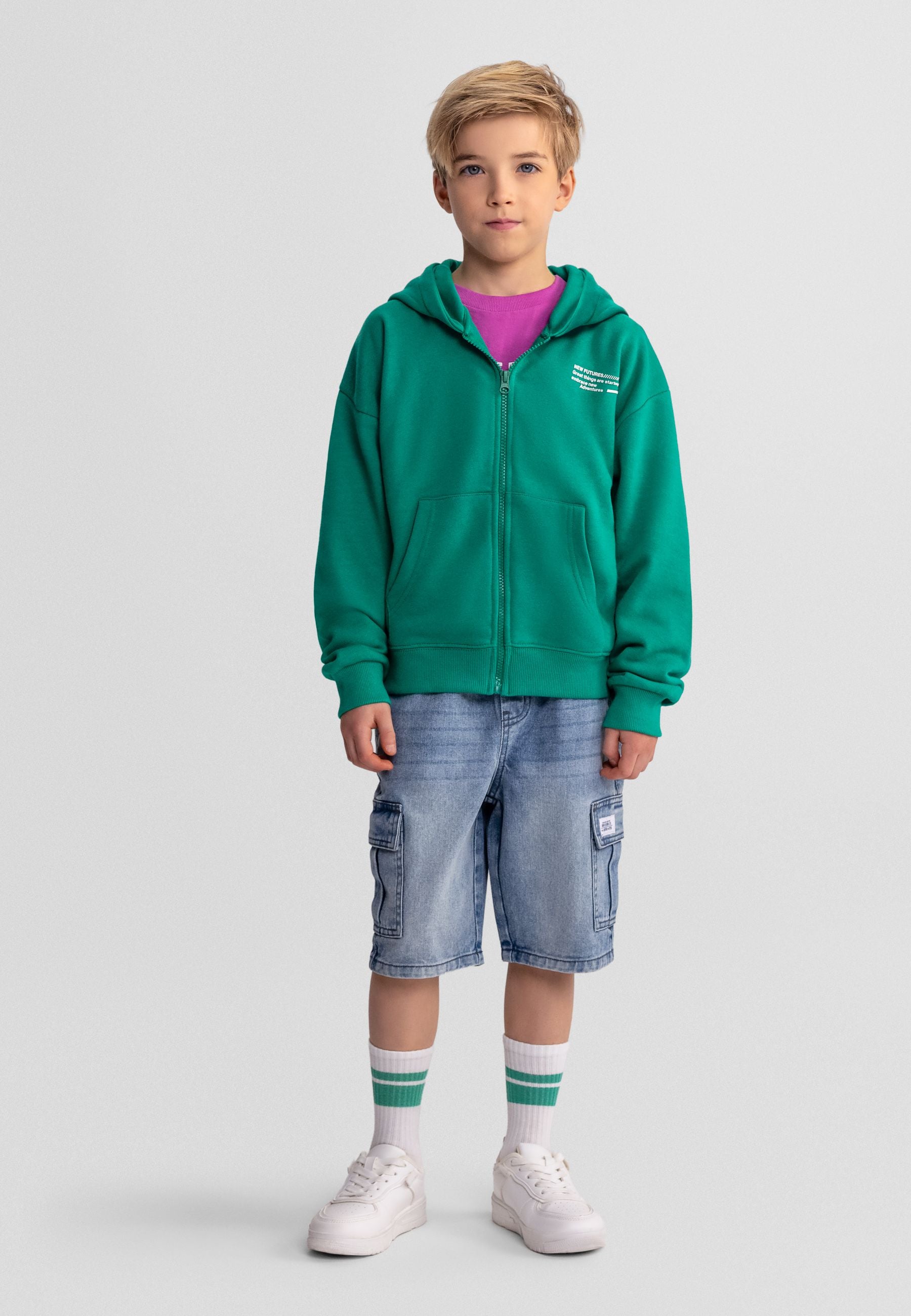 Young blonde boy wearing green zip-up hoodie over purple t-shirt with light blue denim cargo shorts and white-green striped socks, standing against light grey background