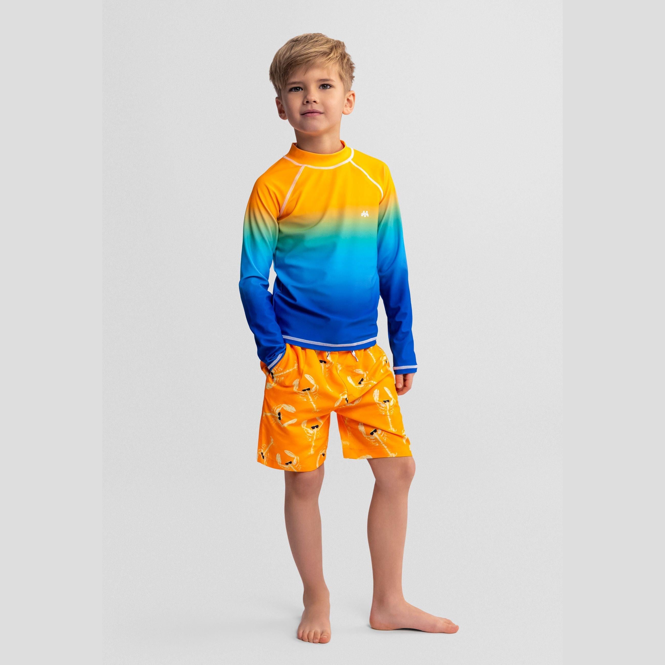 Young boy wearing gradient orange to blue rashguard with orange lobster print swim shorts, standing barefoot against light background