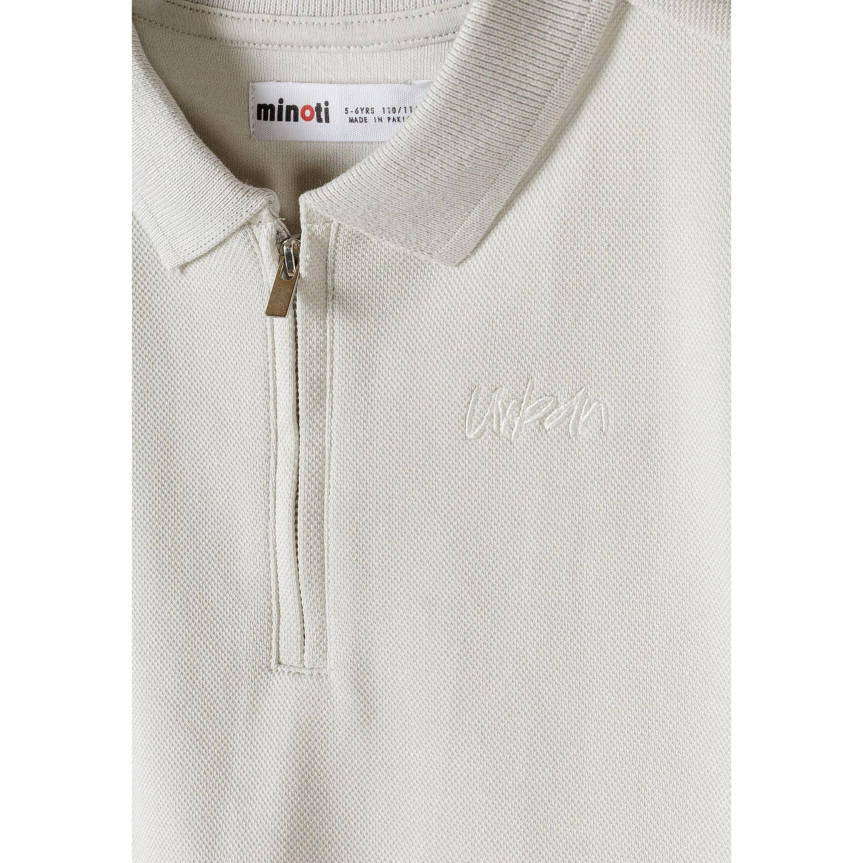 Close-up detail of grey pique polo shirt showing Minoti brand label, zip collar fastening and embroidered logo on textured fabric