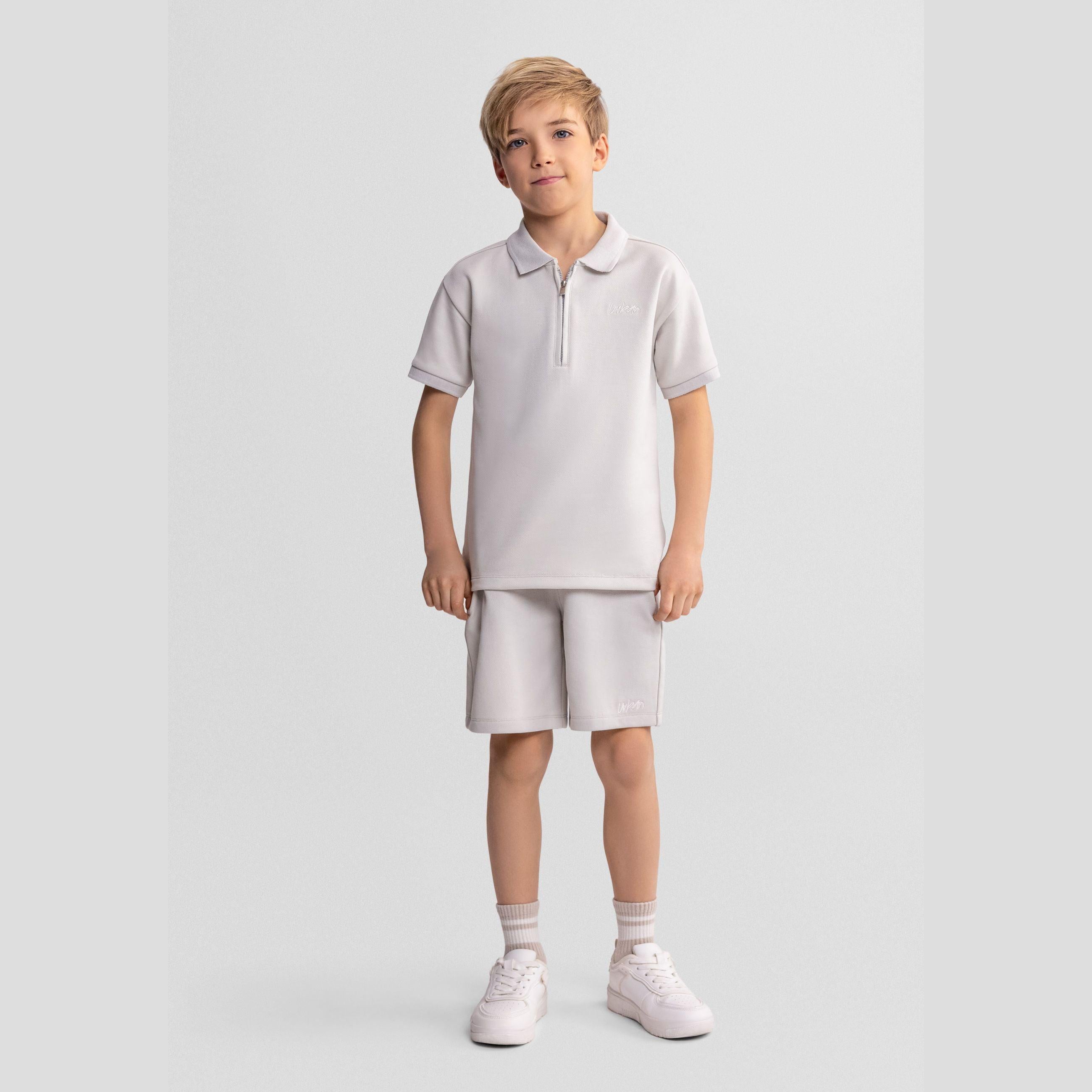 Young boy wearing grey pique polo shirt with zip collar and matching shorts, white trainers and socks, standing against light grey studio background