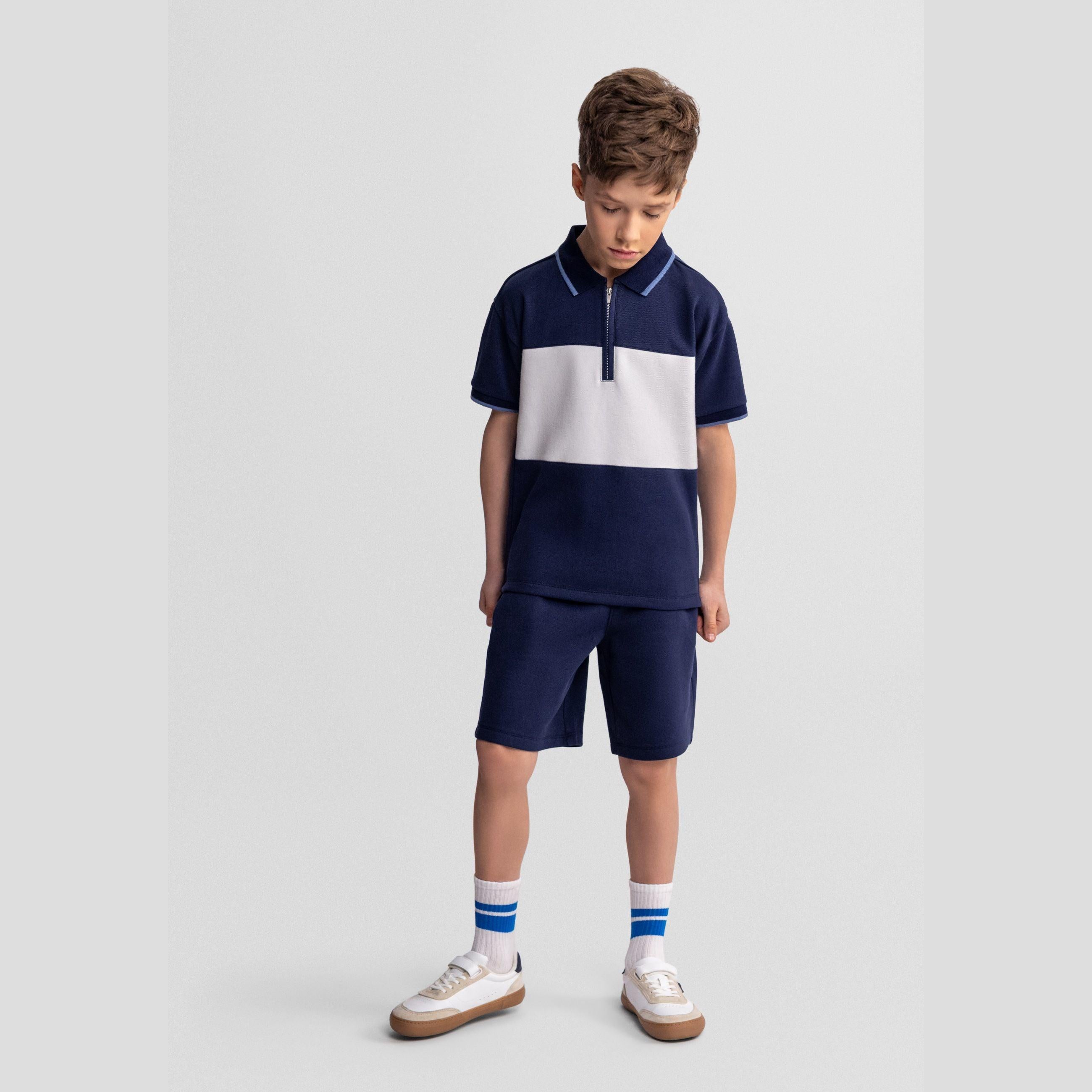 Boy wearing navy and white pique polo set with zip collar and matching shorts, standing against light grey background