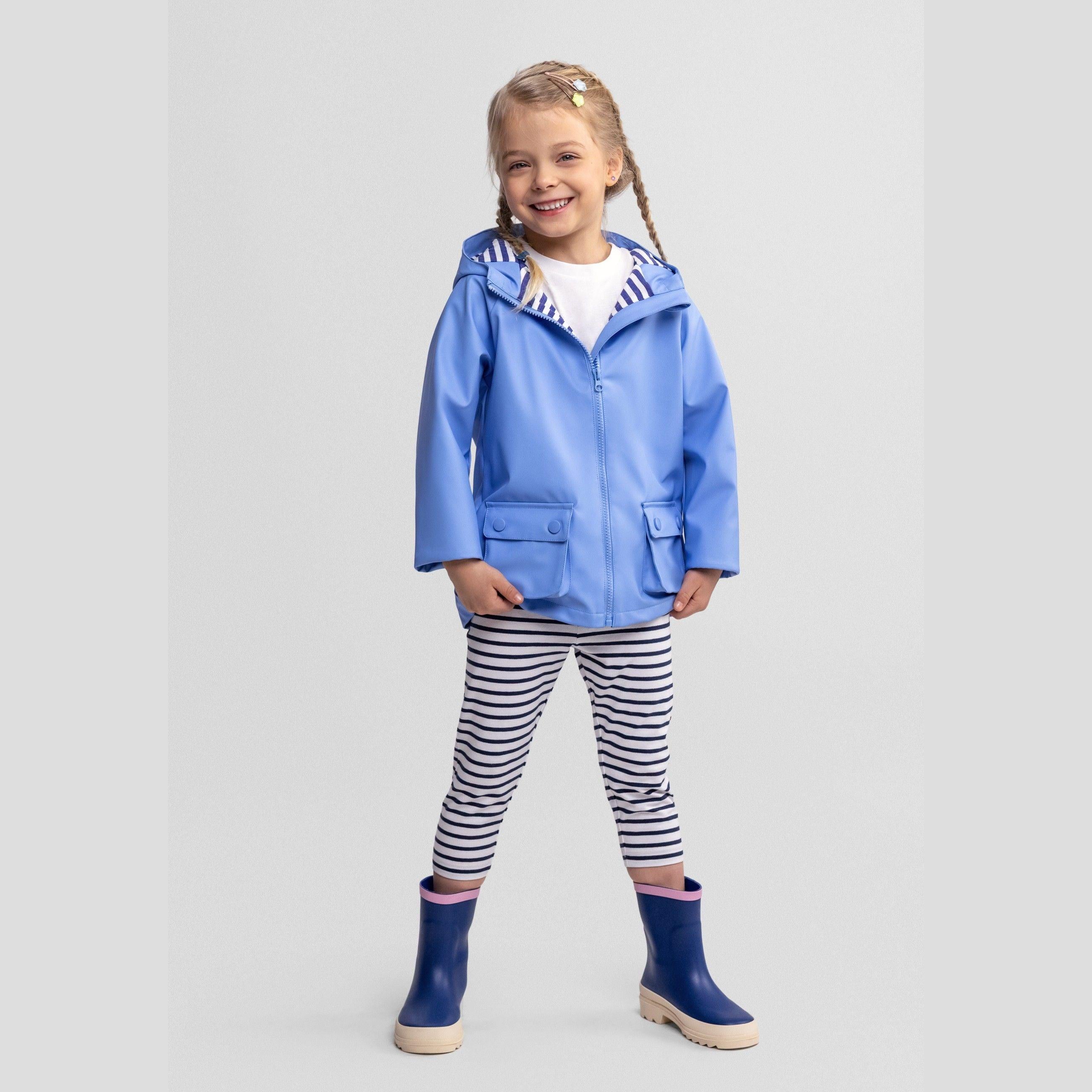 Young girl wearing blue waterproof rain jacket with fixed hood, striped leggings and blue wellington boots on grey background