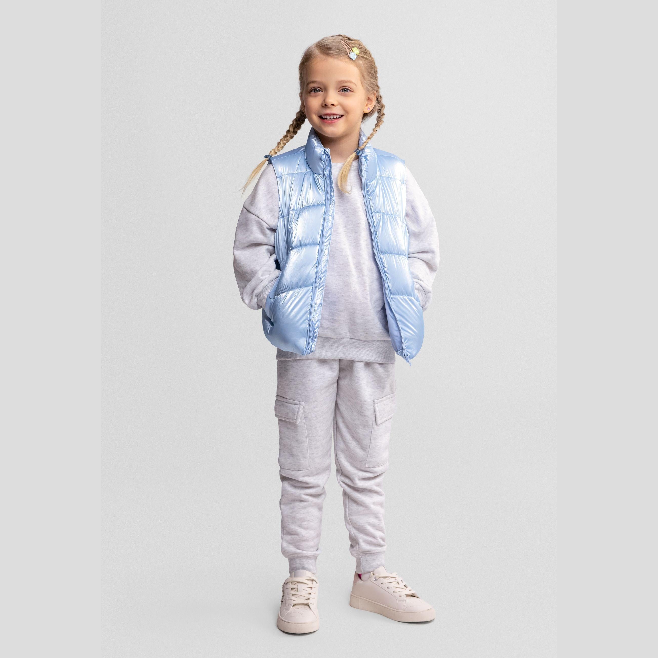 Young girl wearing metallic blue quilted waistcoat over grey hoodie and joggers, standing against light grey background with braided hair and small hair accessory