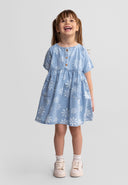 Smiling young girl wearing light blue chambray dress with white daisy print, short sleeves, wooden button front, standing against white background