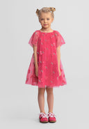 Young blonde girl wearing pink tulle dress with sequin cherry pattern and angel sleeves, standing against white background wearing pink shoes