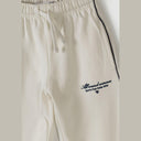 Close-up detail of beige wide-leg tracksuit trousers showing elastic waistband with drawstring and navy embroidered logo