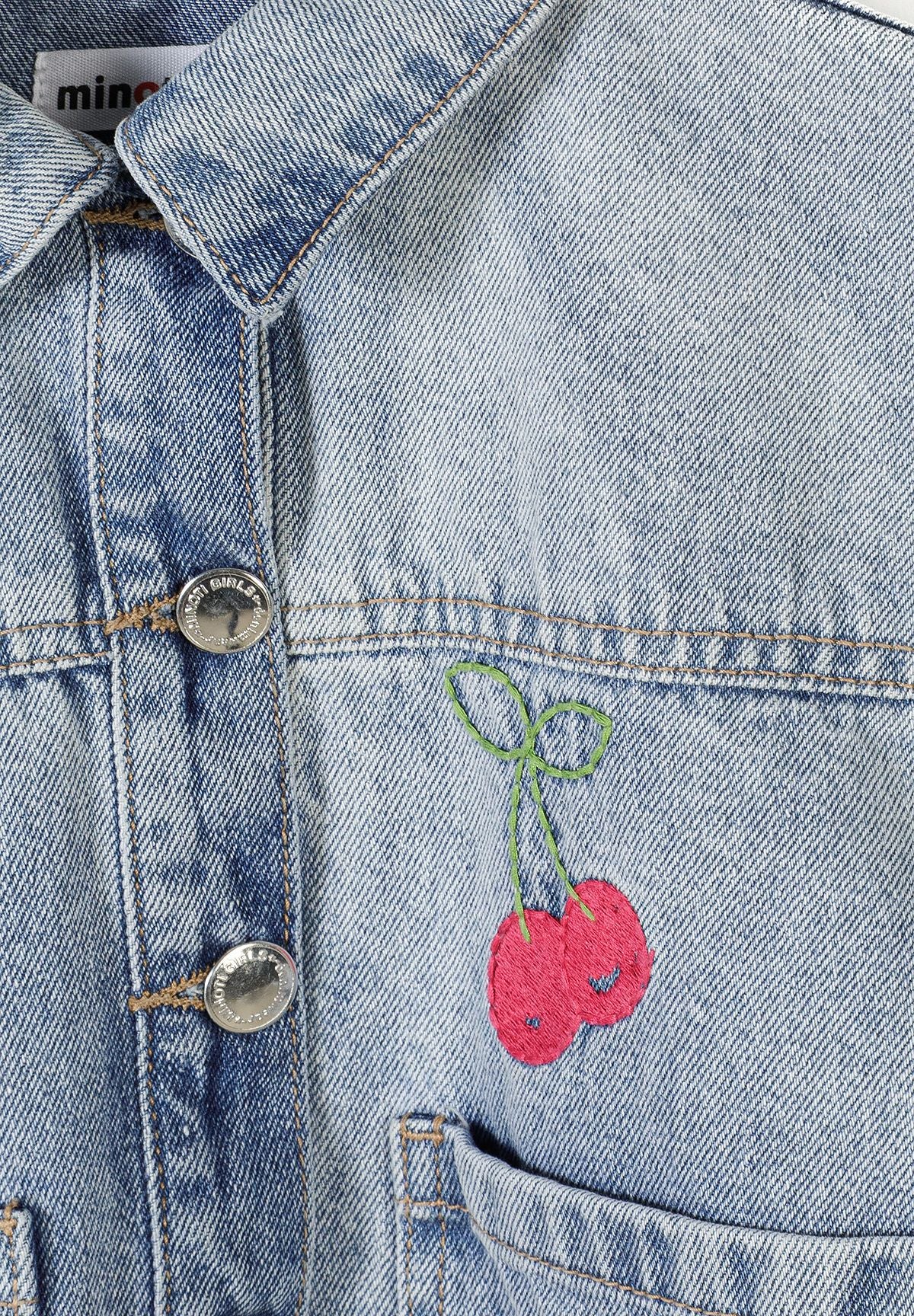 Close-up detail of light blue denim jacket showing embroidered pink cherries with green stems and silver button closure with Minoti branding visible