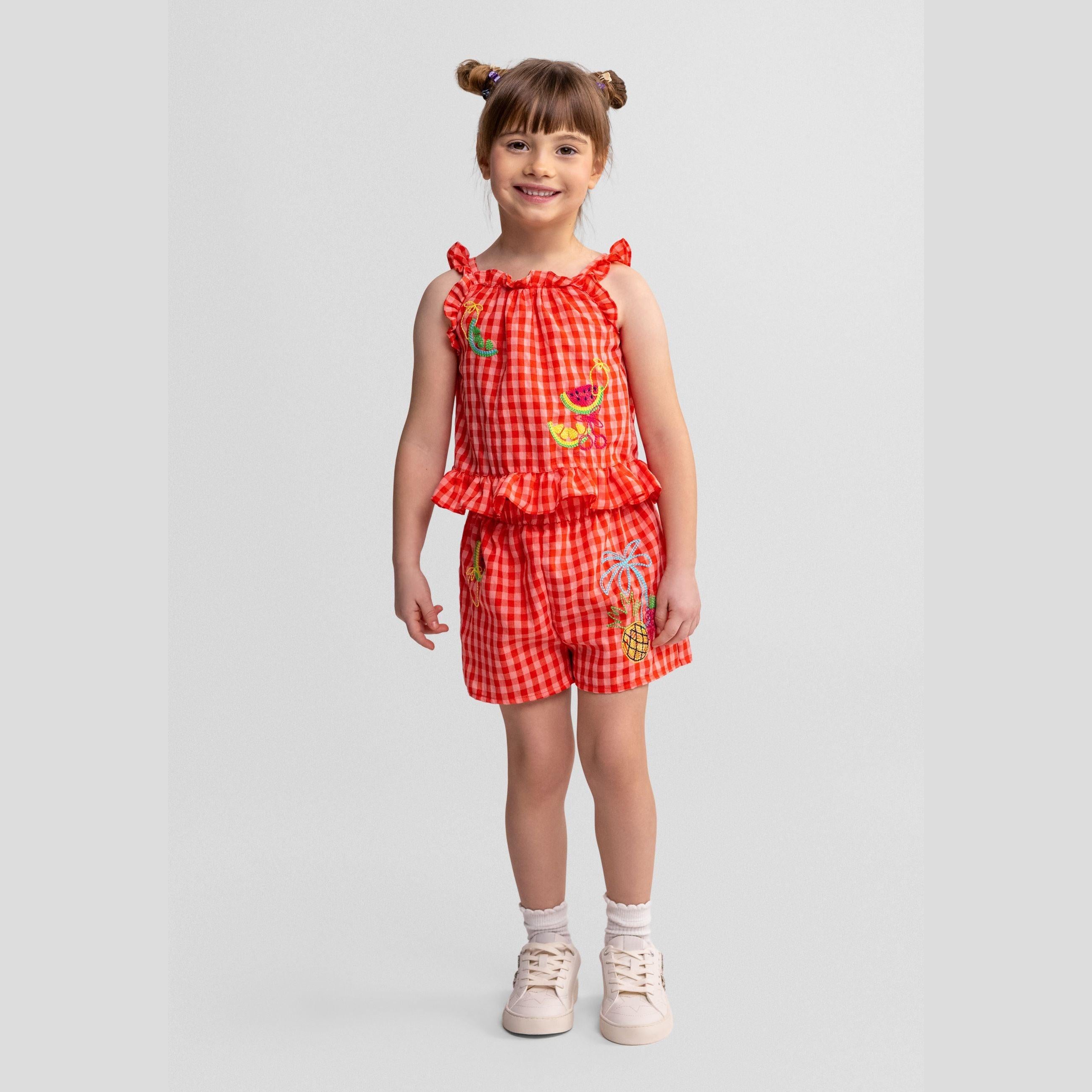 Young girl wearing red and white gingham shorts set with fruit embroidery, ruffle shoulder straps, posed against light grey background