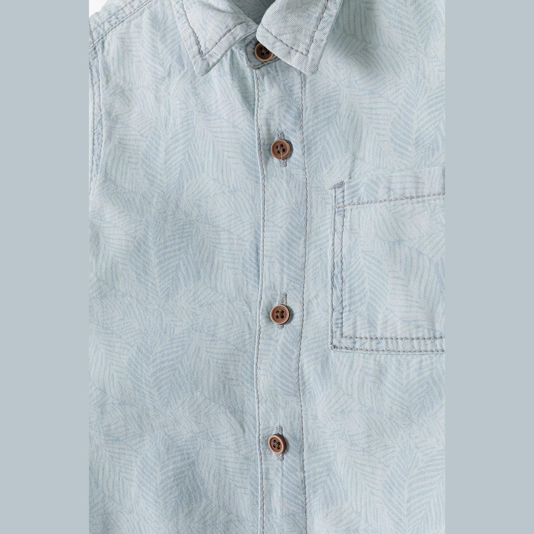 Close-up detail of light blue bleached denim shirt showing wooden-effect buttons, chest pocket stitching and collar construction