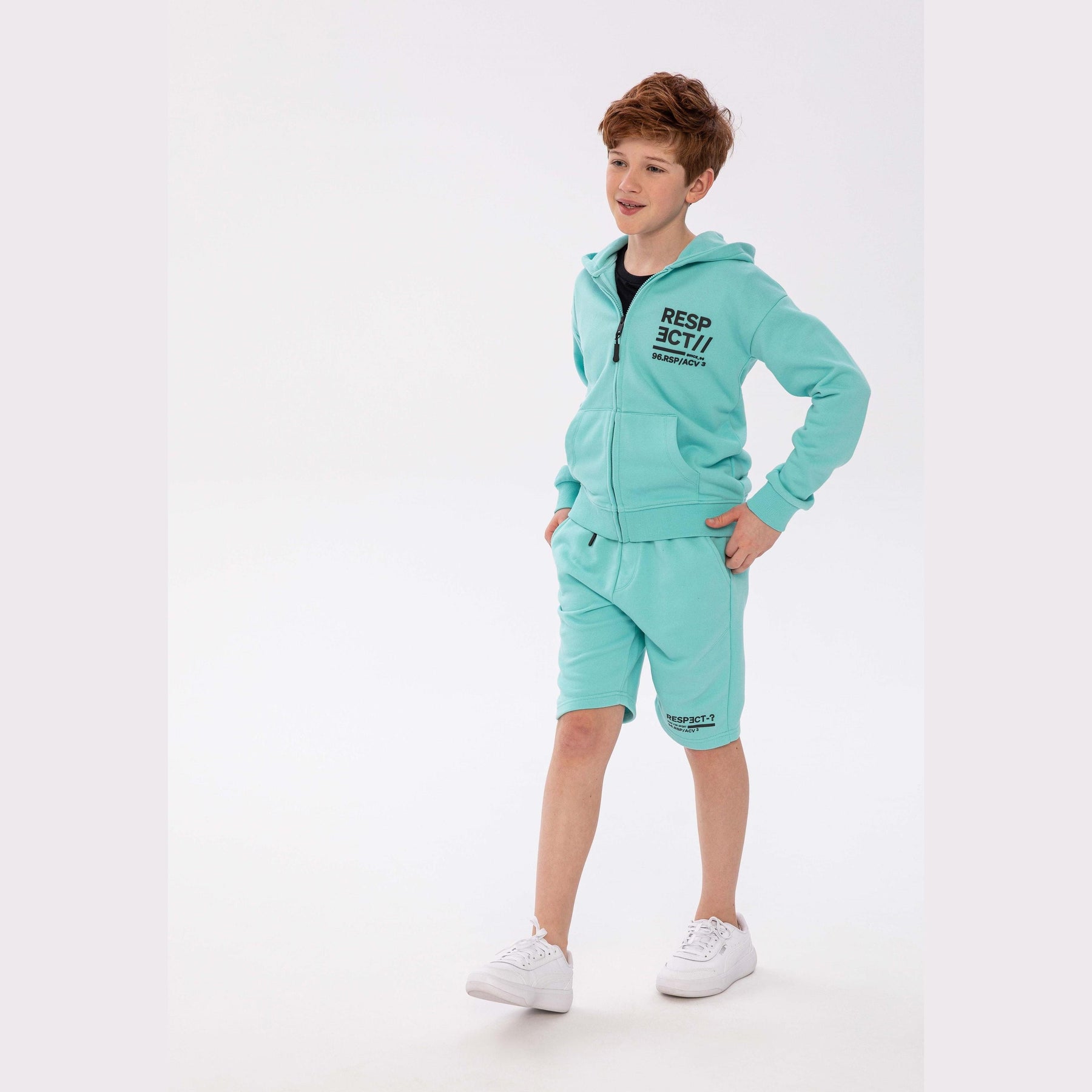 Boy wearing turquoise fleece shorts with matching hoodie, standing on white background with hands on hips, showing complete casual outfit
