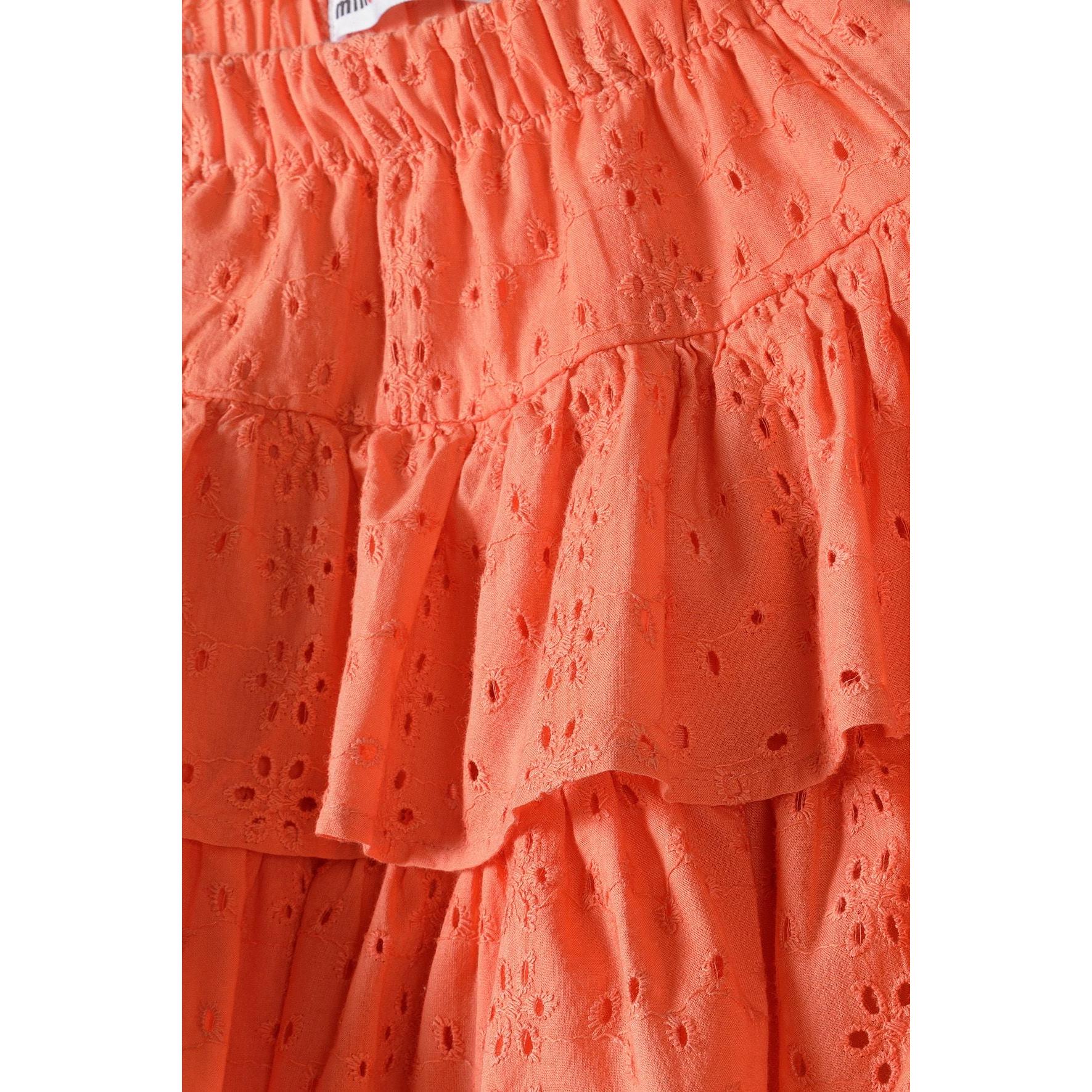 Close-up detail of coral broderie anglaise fabric showing intricate embroidered pattern and ruffle construction with Minoti label visible