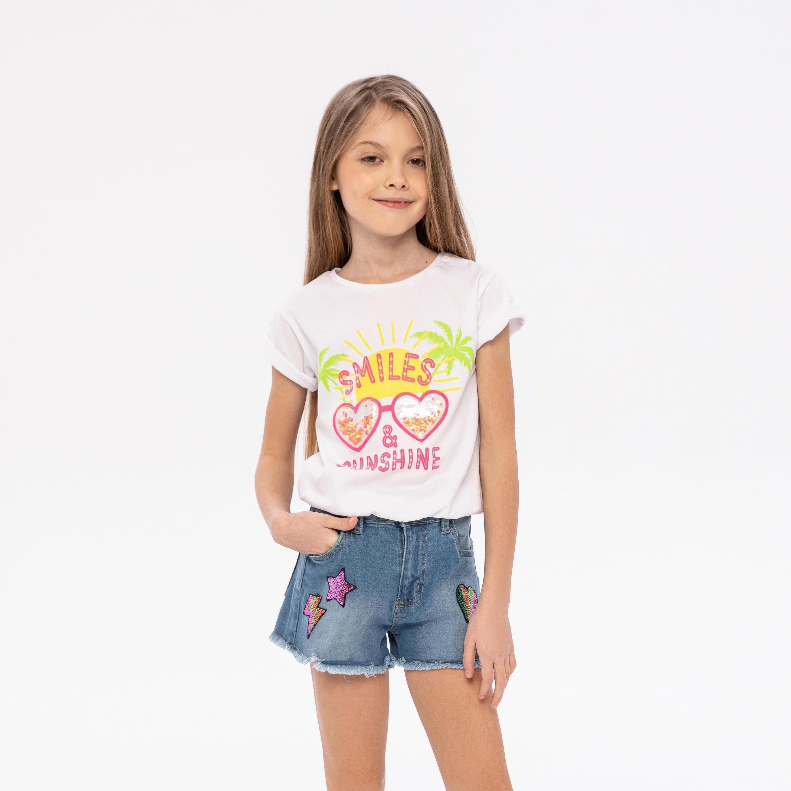 Young girl wearing white t-shirt with smile and sunshine print and denim shorts, standing against white background