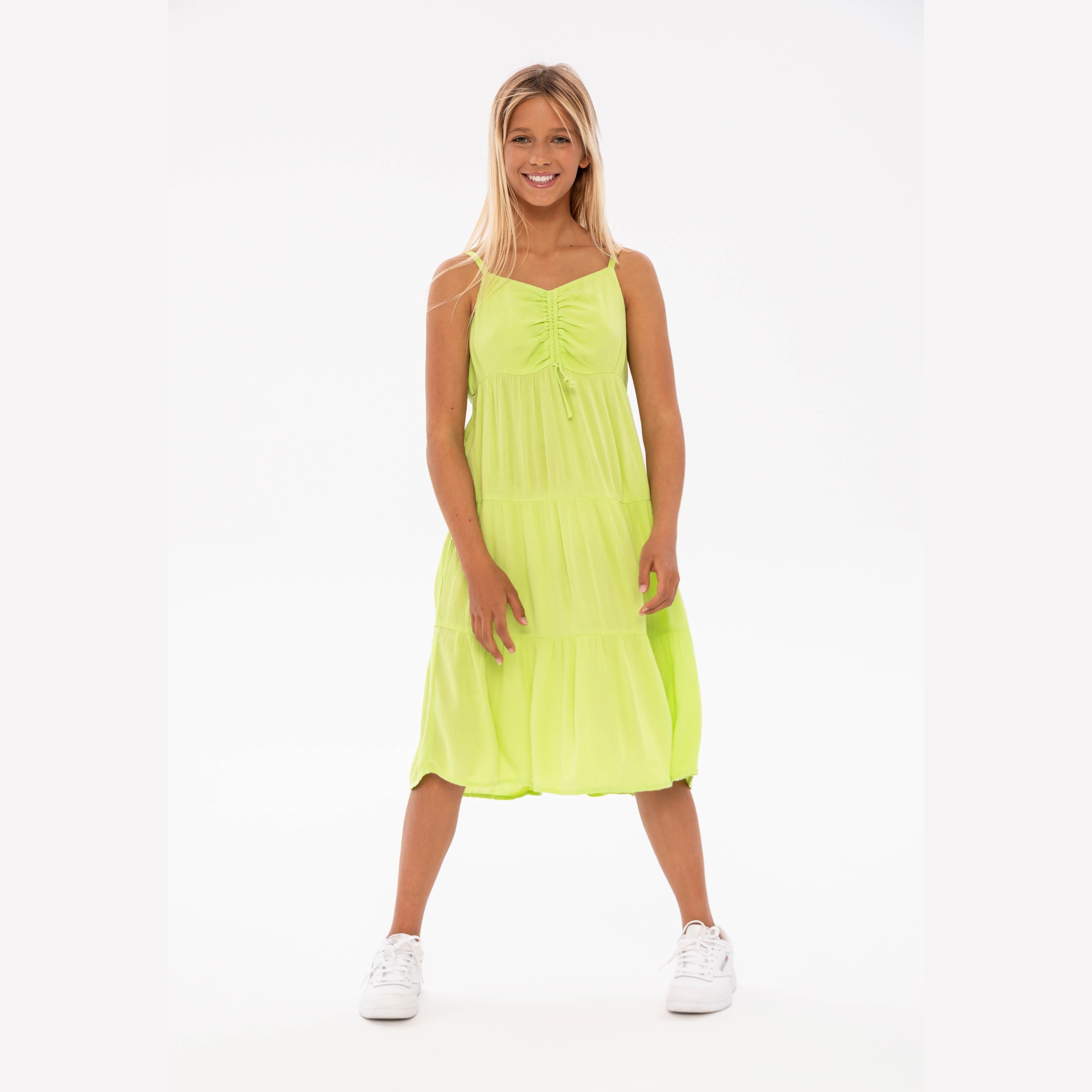 Young woman modeling bright green tiered strappy dress with white sneakers against light background, full body front view
