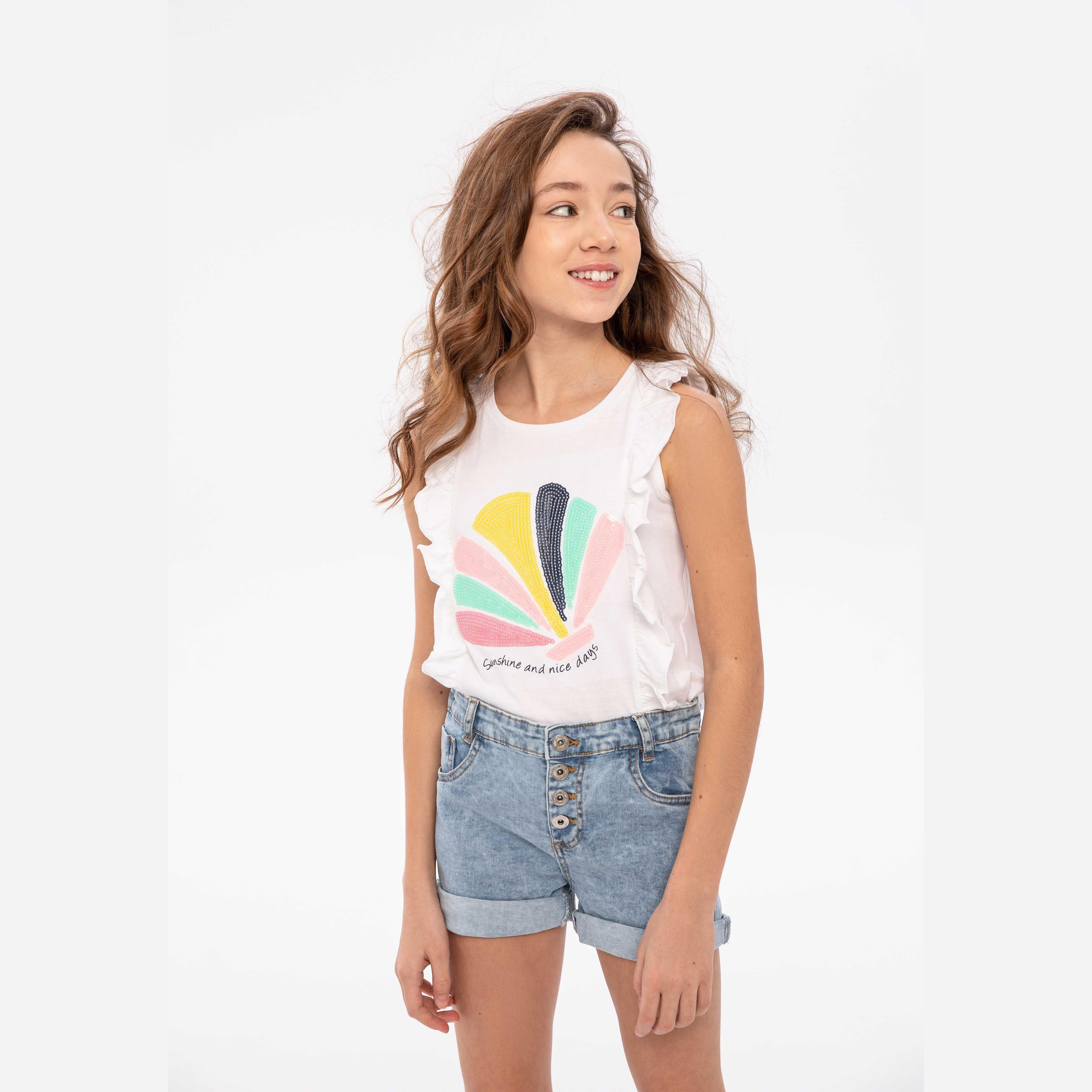 Young girl wearing light blue denim shorts with sequin pocket and white graphic t-shirt, standing against white background, smiling