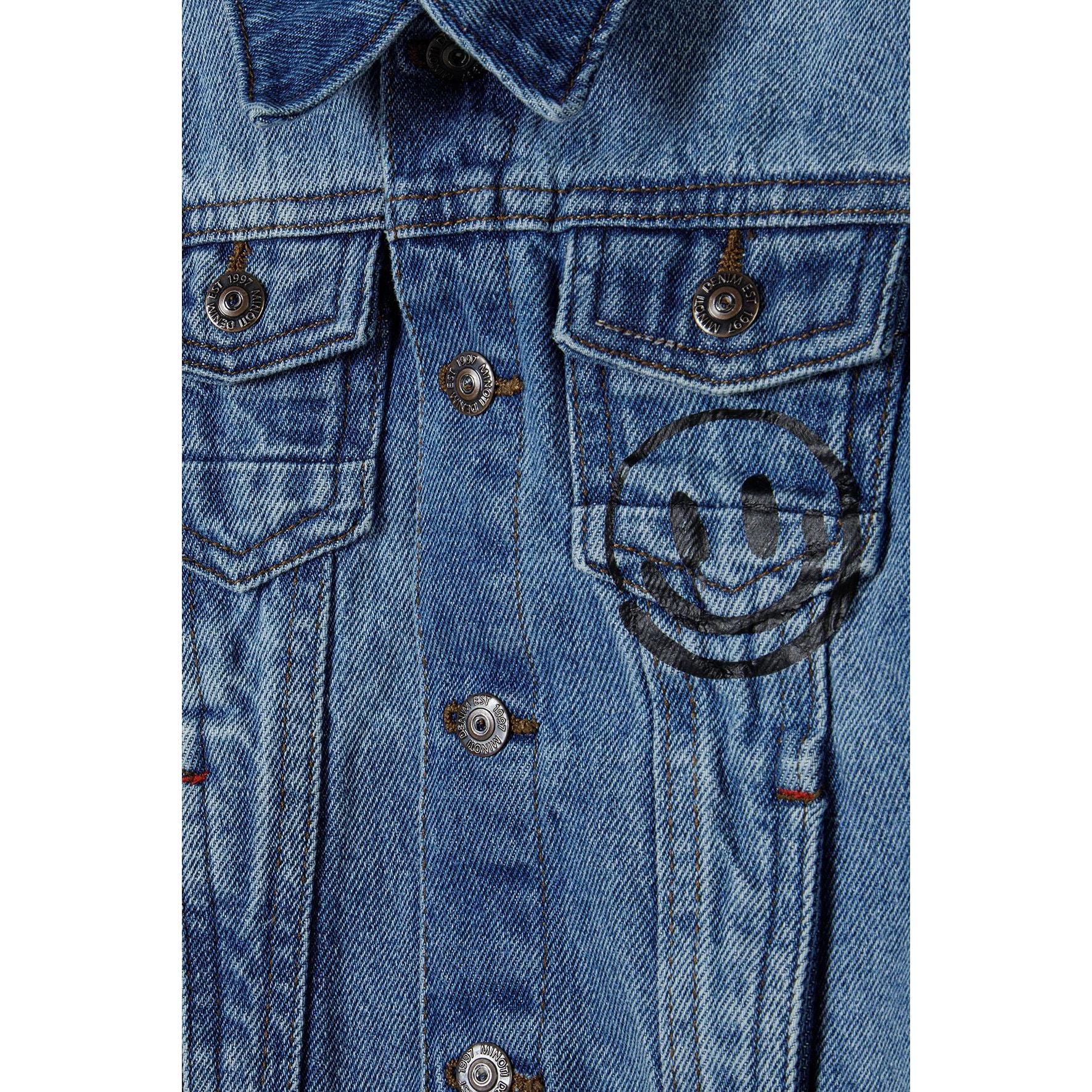 Close-up detail of light blue denim jacket front showing button closure, chest pockets, circular badge patch, and classic denim stitching details