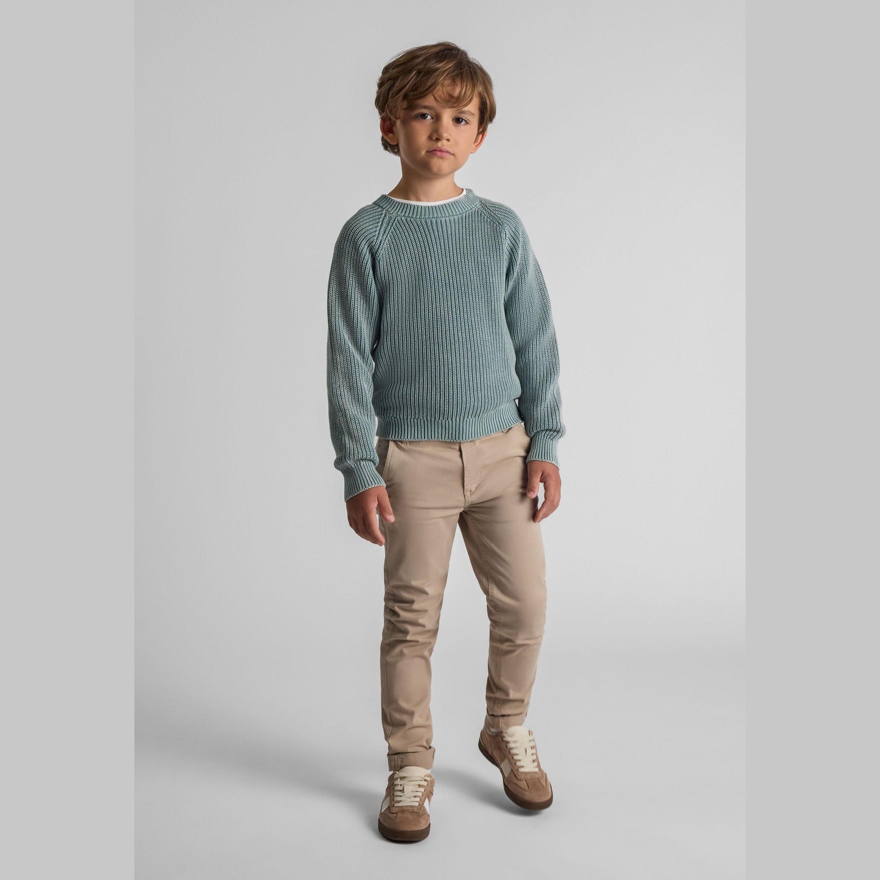 Young boy wearing grey chino trousers with sage green knitted sweater and beige trainers, standing against white background in studio setting