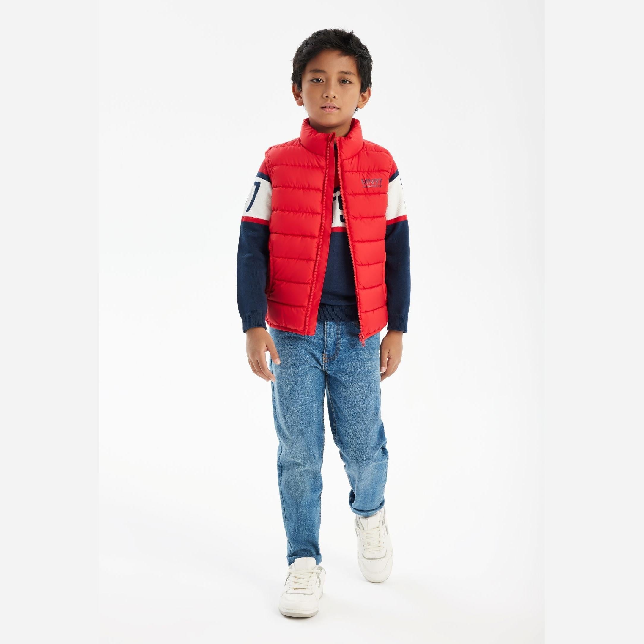 Young boy wearing red padded gilet over navy sweatshirt with jeans and white trainers, standing against white background