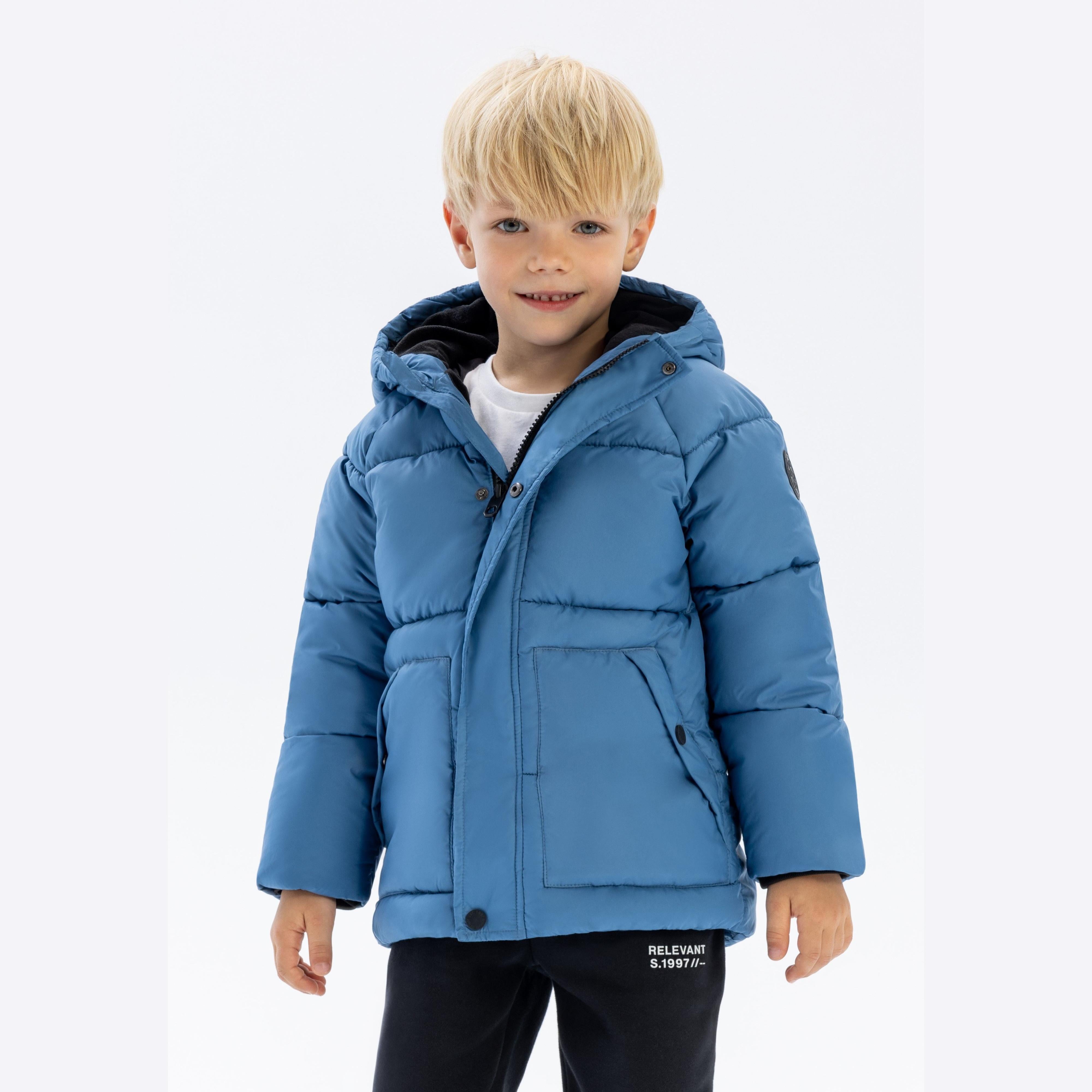 Young blonde boy wearing bright blue padded jacket with hood up, smiling at camera against white background, paired with dark trousers