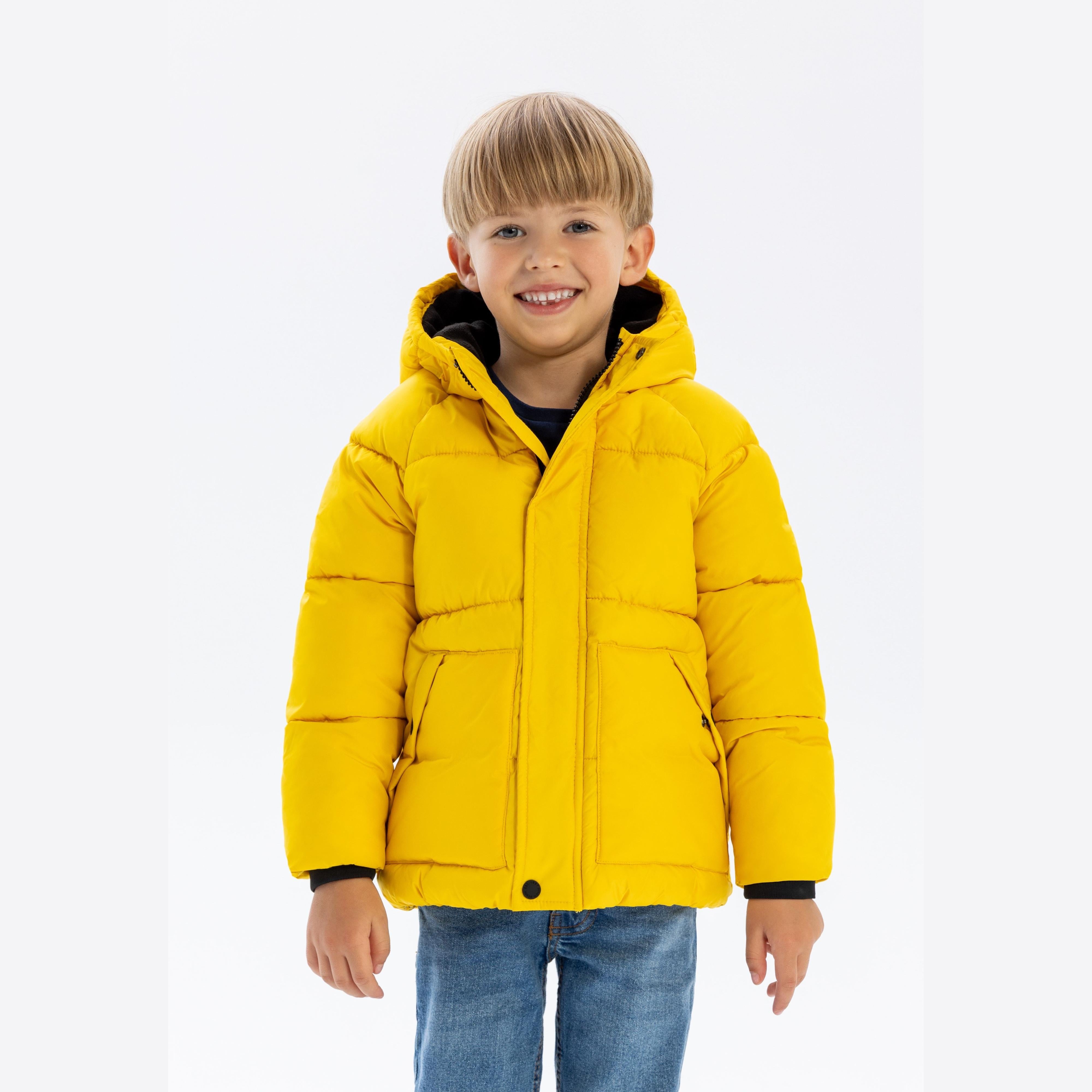 Young blonde boy wearing bright yellow padded jacket with hood and dark cuffs, paired with blue jeans, smiling at camera against white studio background
