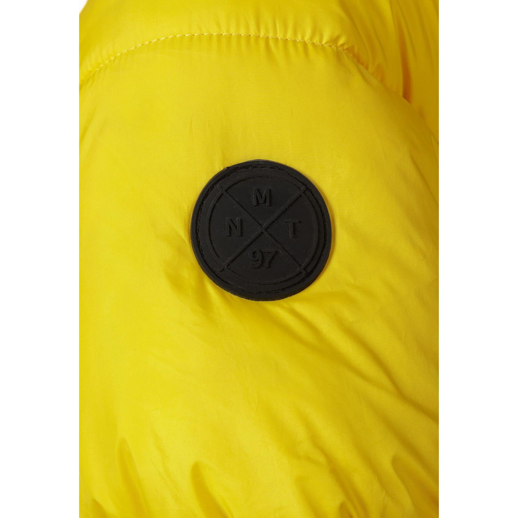 Close-up detail of black circular Minoti logo patch with crossed design on bright yellow padded jacket fabric showing quilted texture