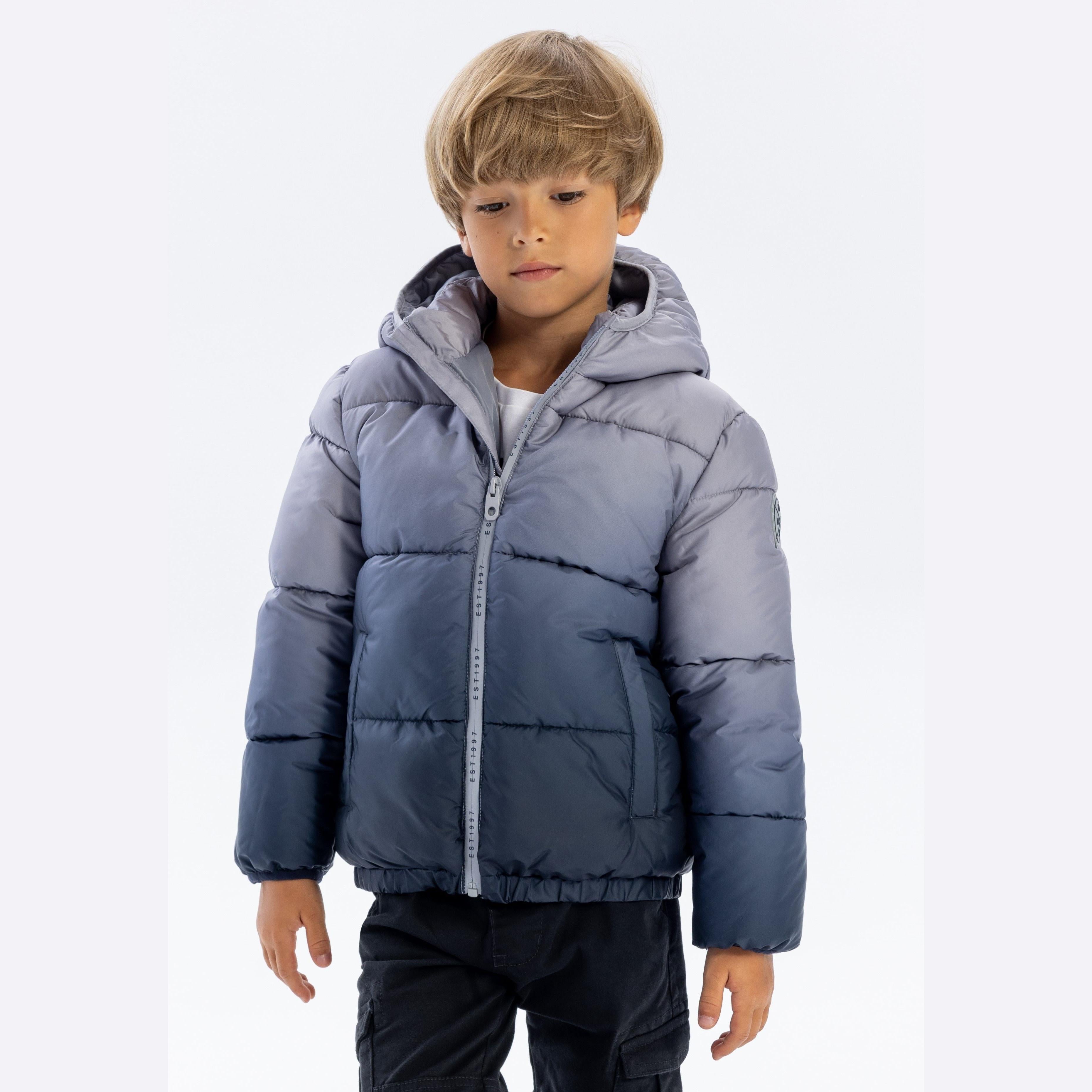Young boy wearing grey ombré padded jacket with hood up, hands at sides, against white background showing full front view of winter coat with zip closure