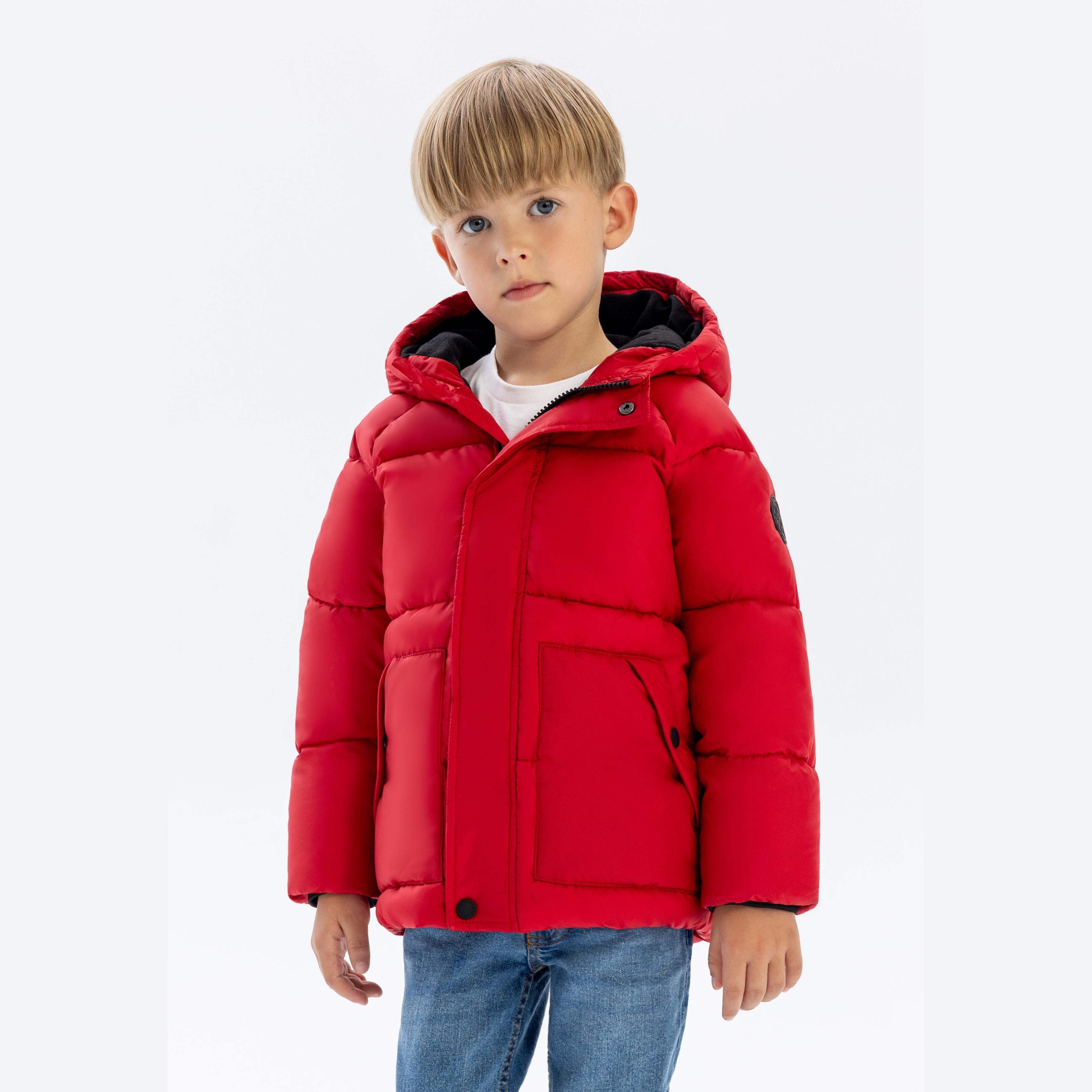 Young blonde boy wearing bright red padded winter jacket with hood and blue jeans, standing against white studio background