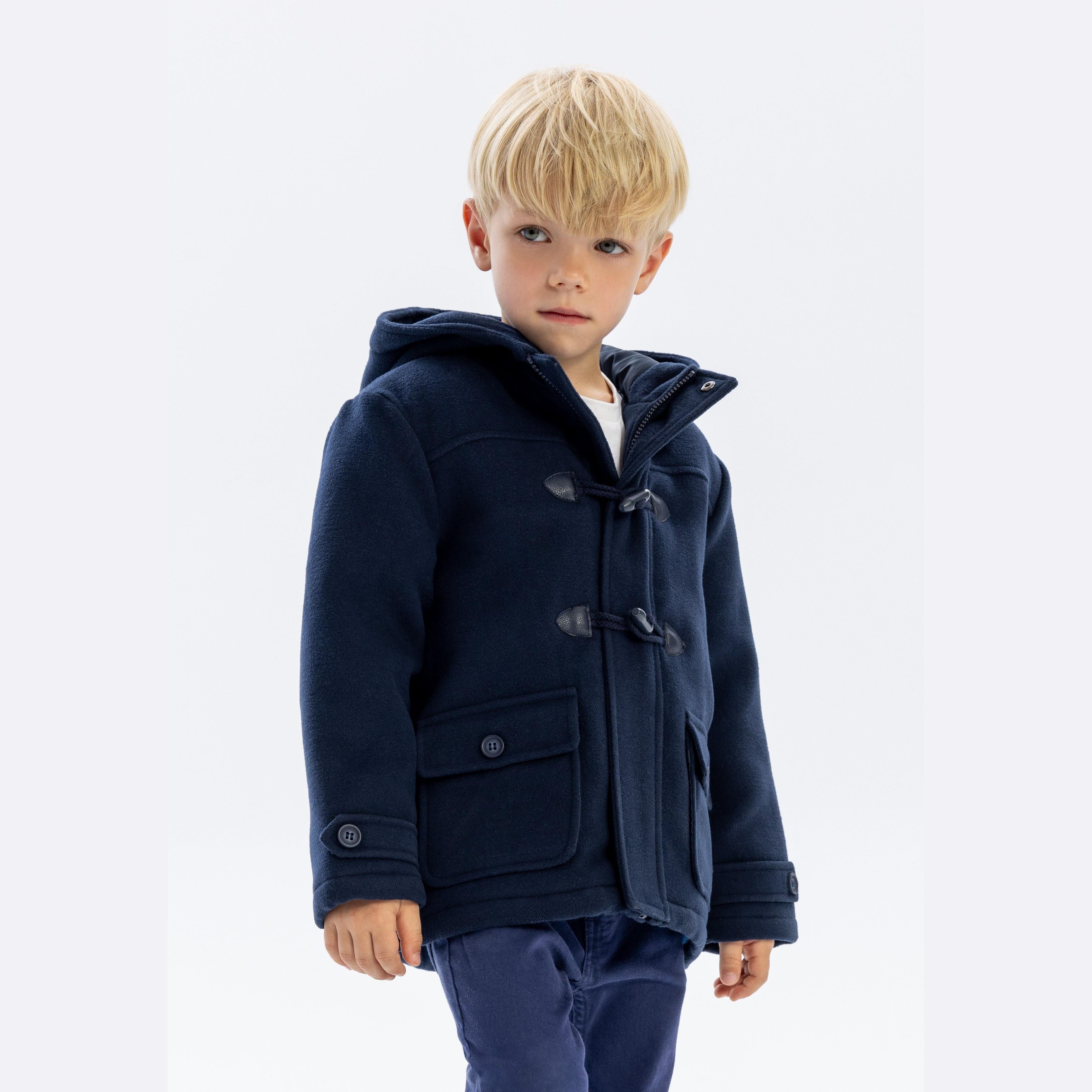Young blonde boy wearing navy padded duffle coat with hood and toggle fastenings, standing against white background