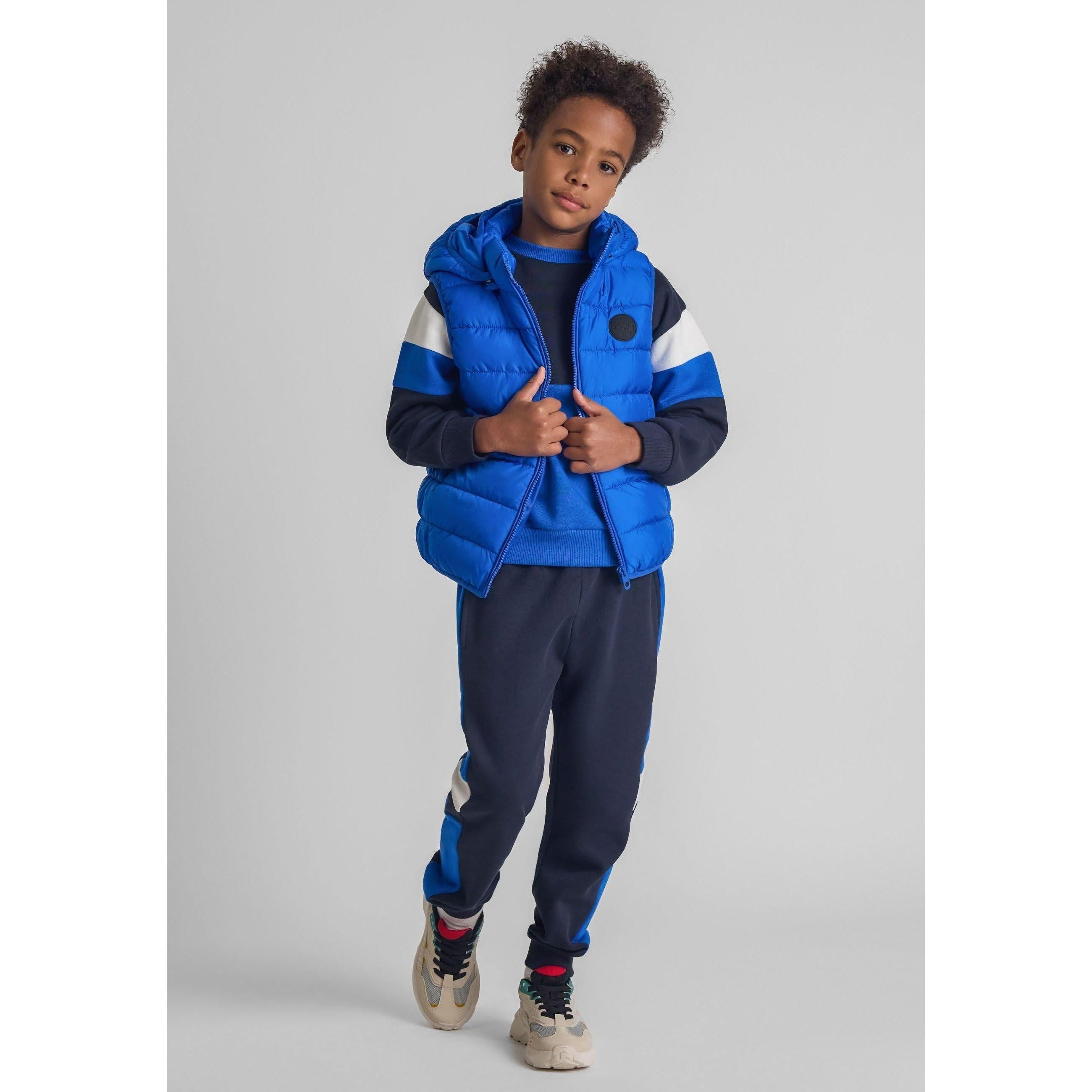 Boy wearing cobalt blue padded gilet with detachable hood over navy tracksuit, standing against grey background