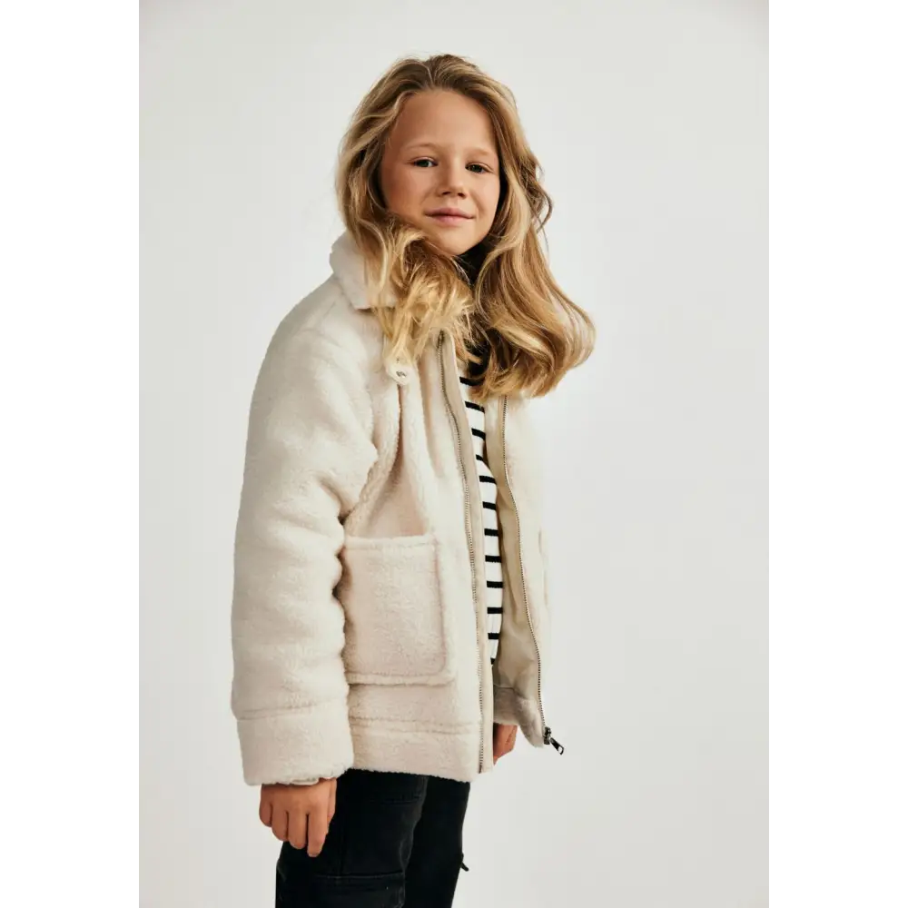 Young girl wearing cream teddy borg jacket with hood up over striped top and dark jeans, standing against white background