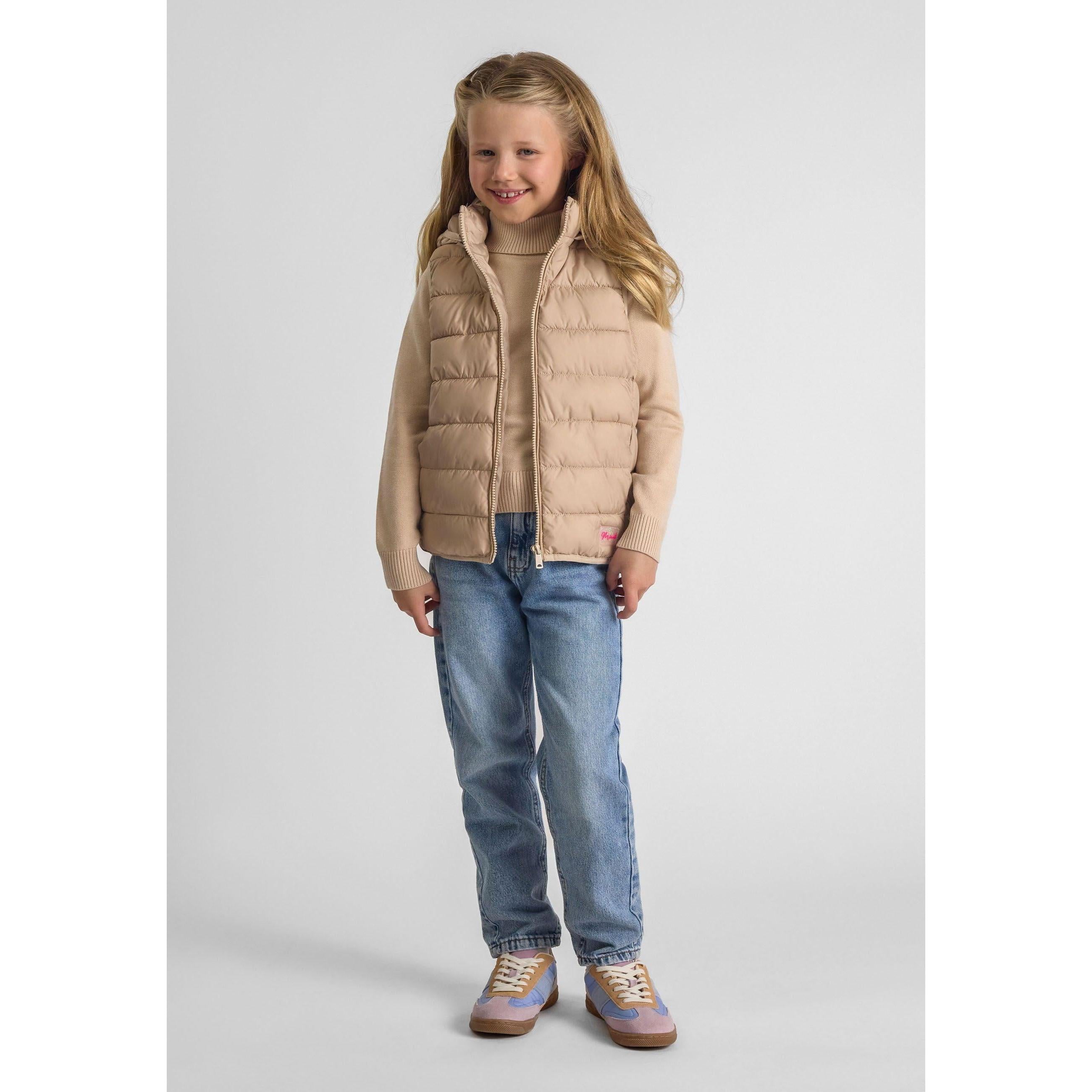 Young girl wearing beige quilted gilet with detachable hood over knit sweater and jeans, standing against white background