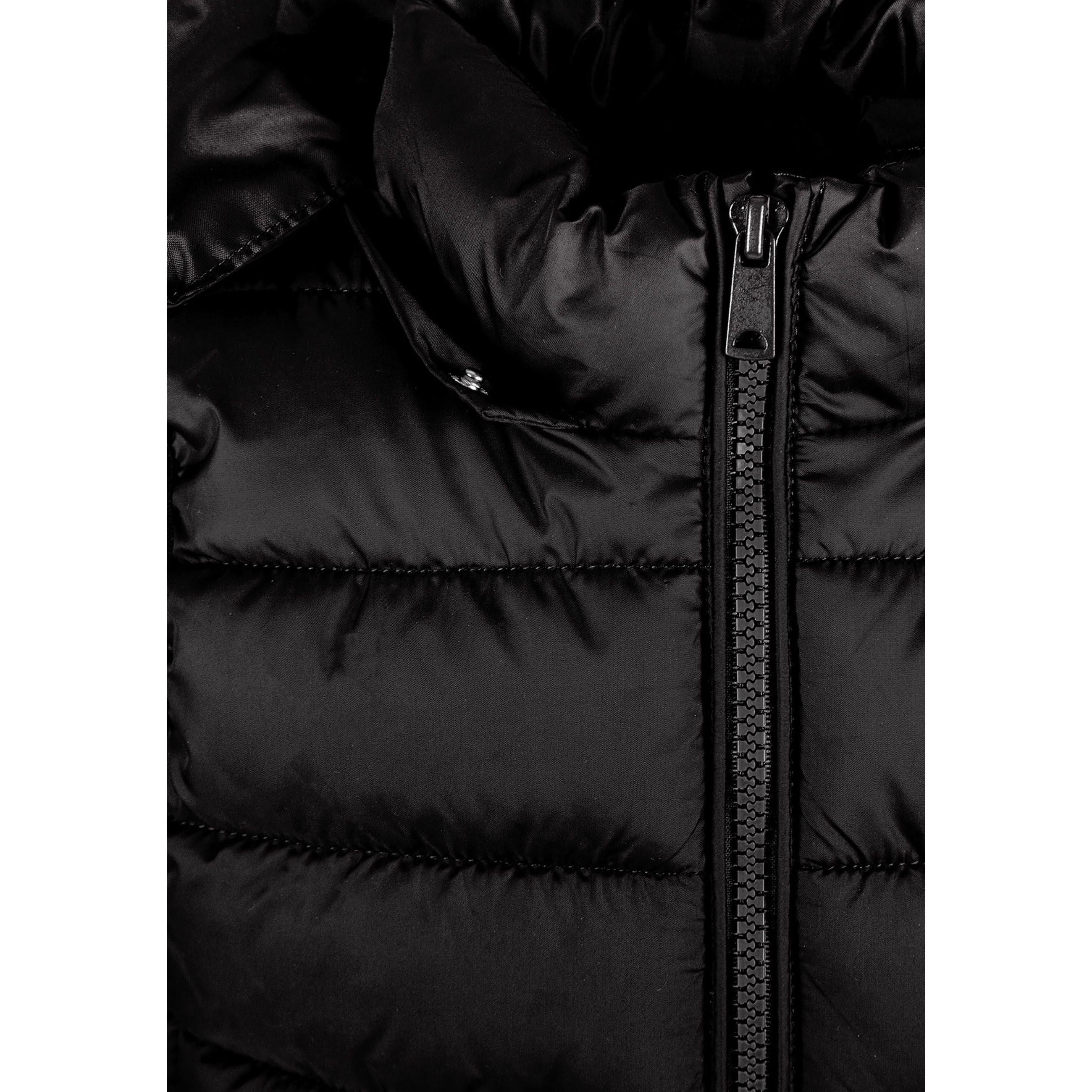 Girl in black quilted gilet zipped up over white hoodie and cream tracksuit, hands positioned on zip, against white studio background