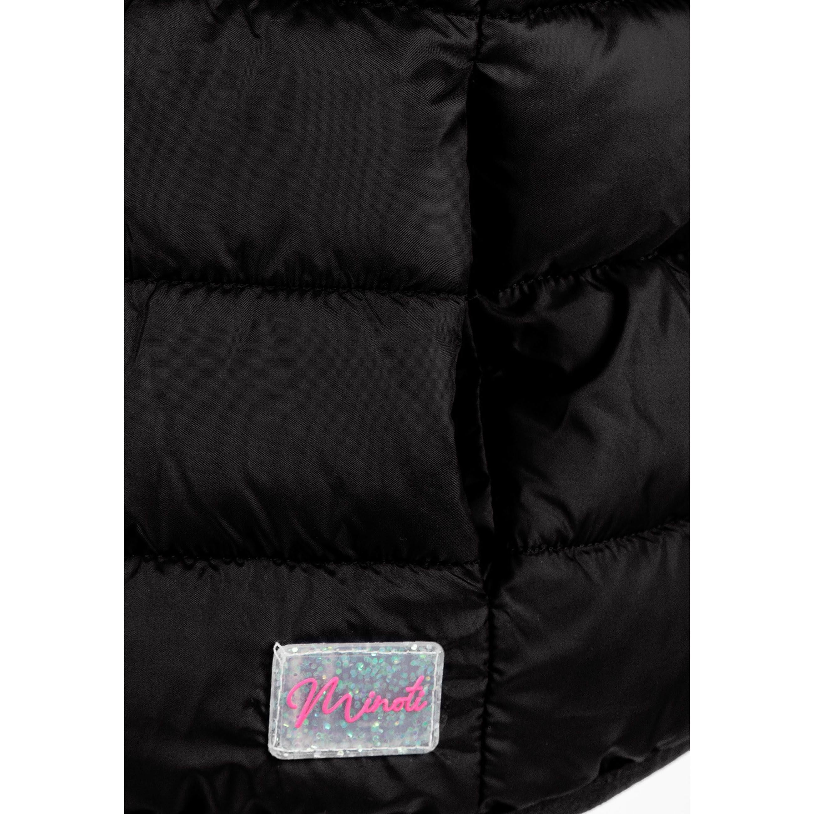 Girl in black quilted gilet zipped up over white hoodie and cream tracksuit, hands positioned on zip, against white studio background