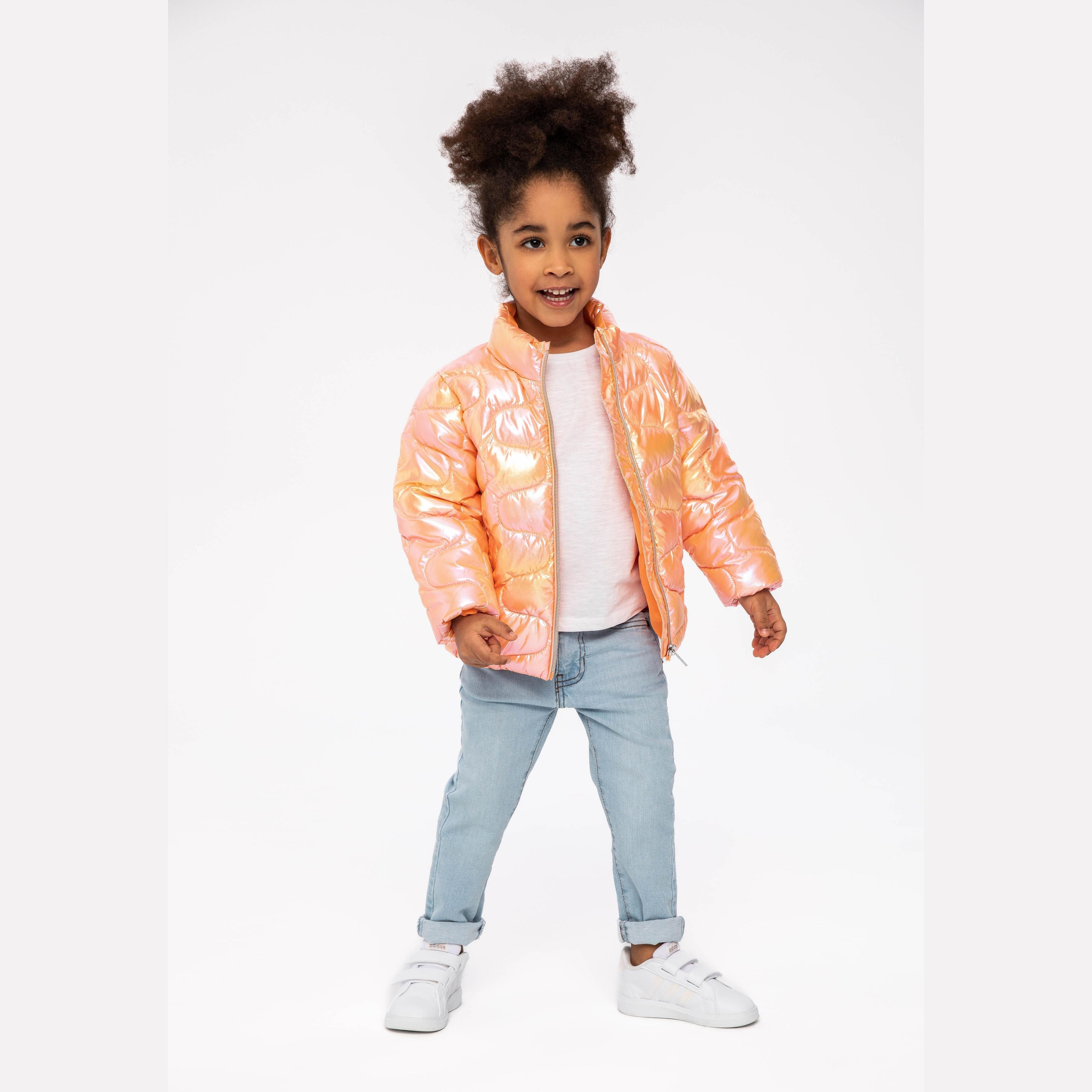 Young girl wearing iridescent coral quilted jacket with white top and light blue jeans, smiling against white background