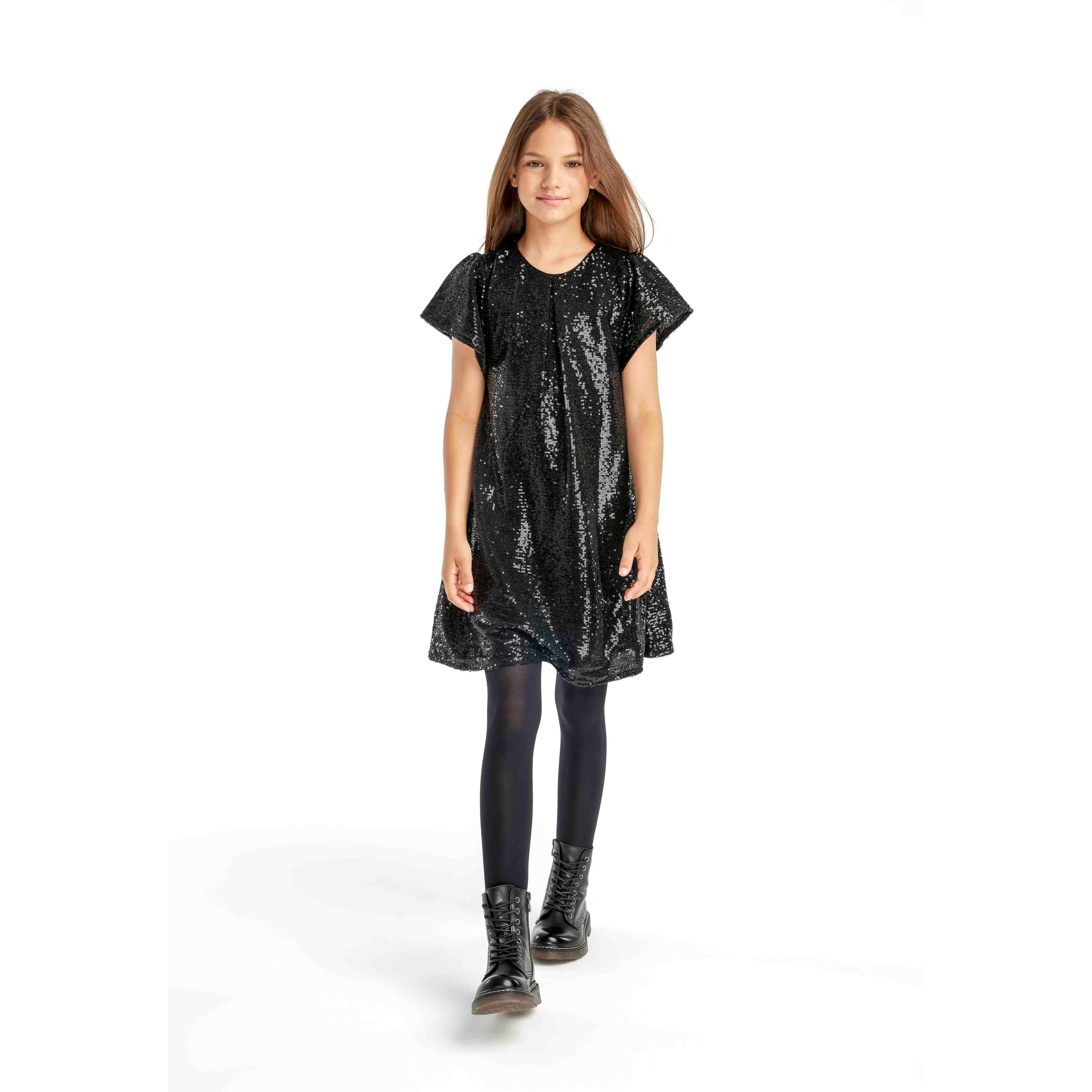 Young girl wearing black sequin party dress with flutter sleeves, paired with black tights and boots, standing against white background