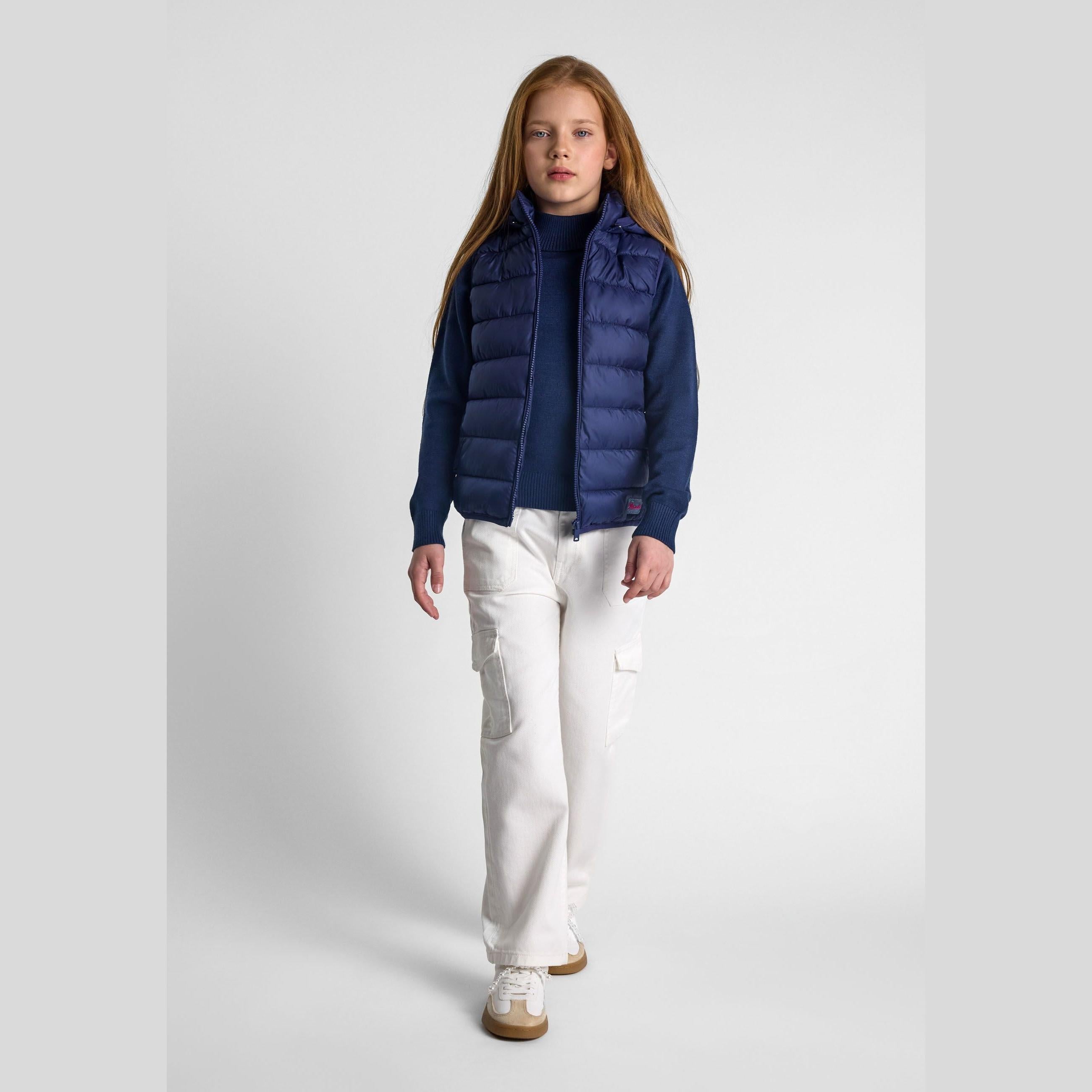 Young girl modeling navy quilted gilet with detachable hood over dark sweater and white trousers, front view on white background