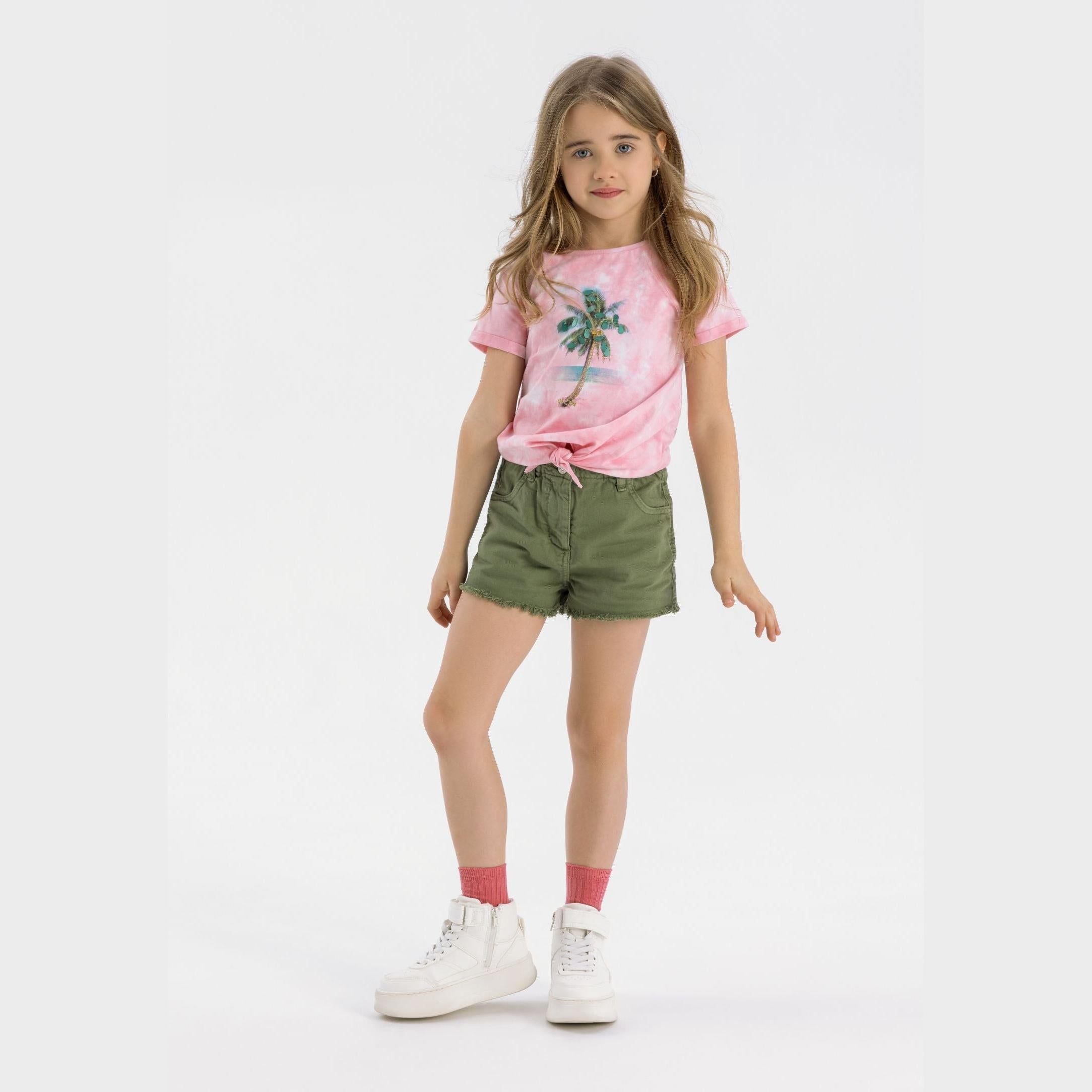 Young girl wearing pink palm tree t-shirt and khaki twill shorts with paperbag waist, coral socks and white sneakers on neutral background