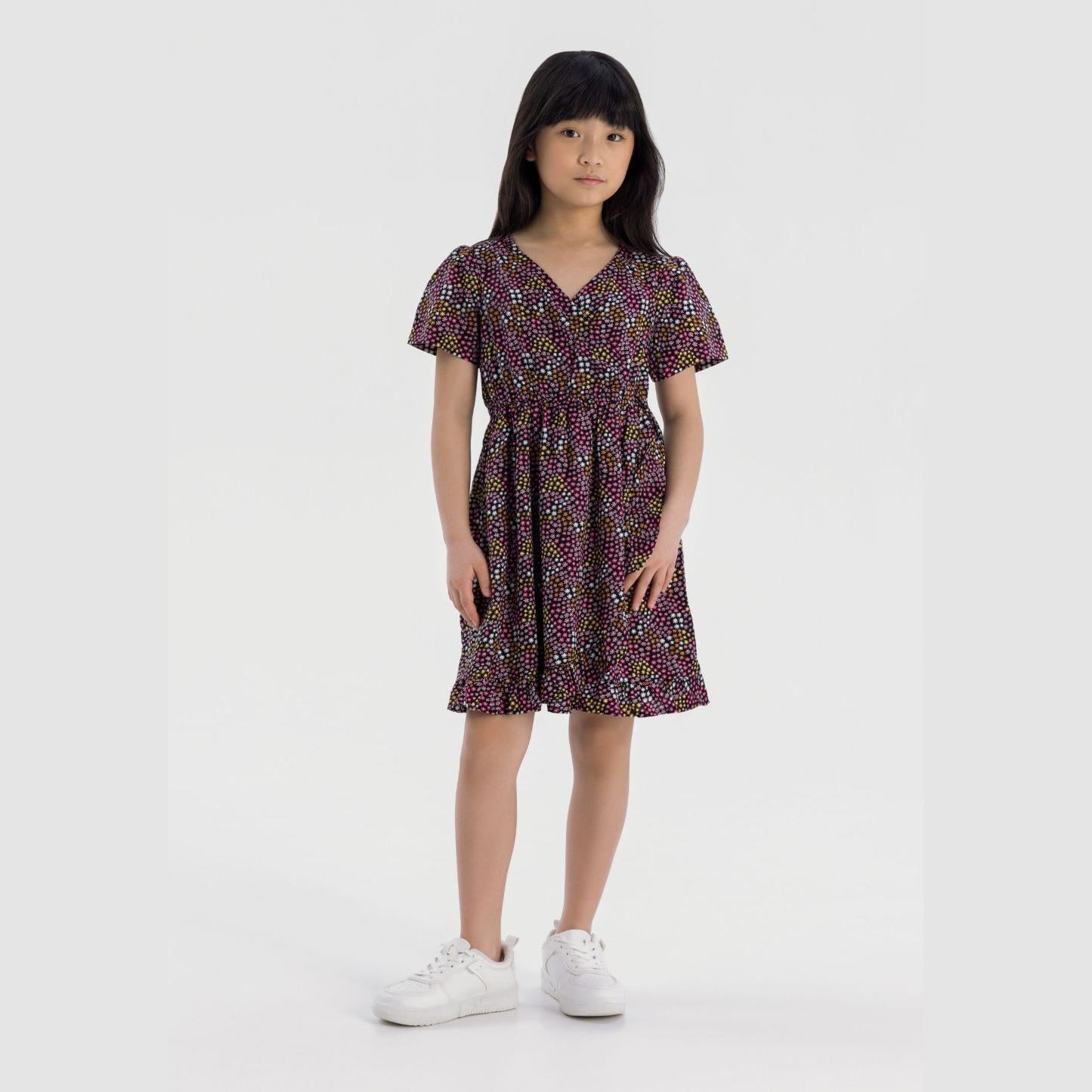 Young girl wearing multicoloured star print wrap dress with short sleeves and frill hem, standing against white background wearing white trainers